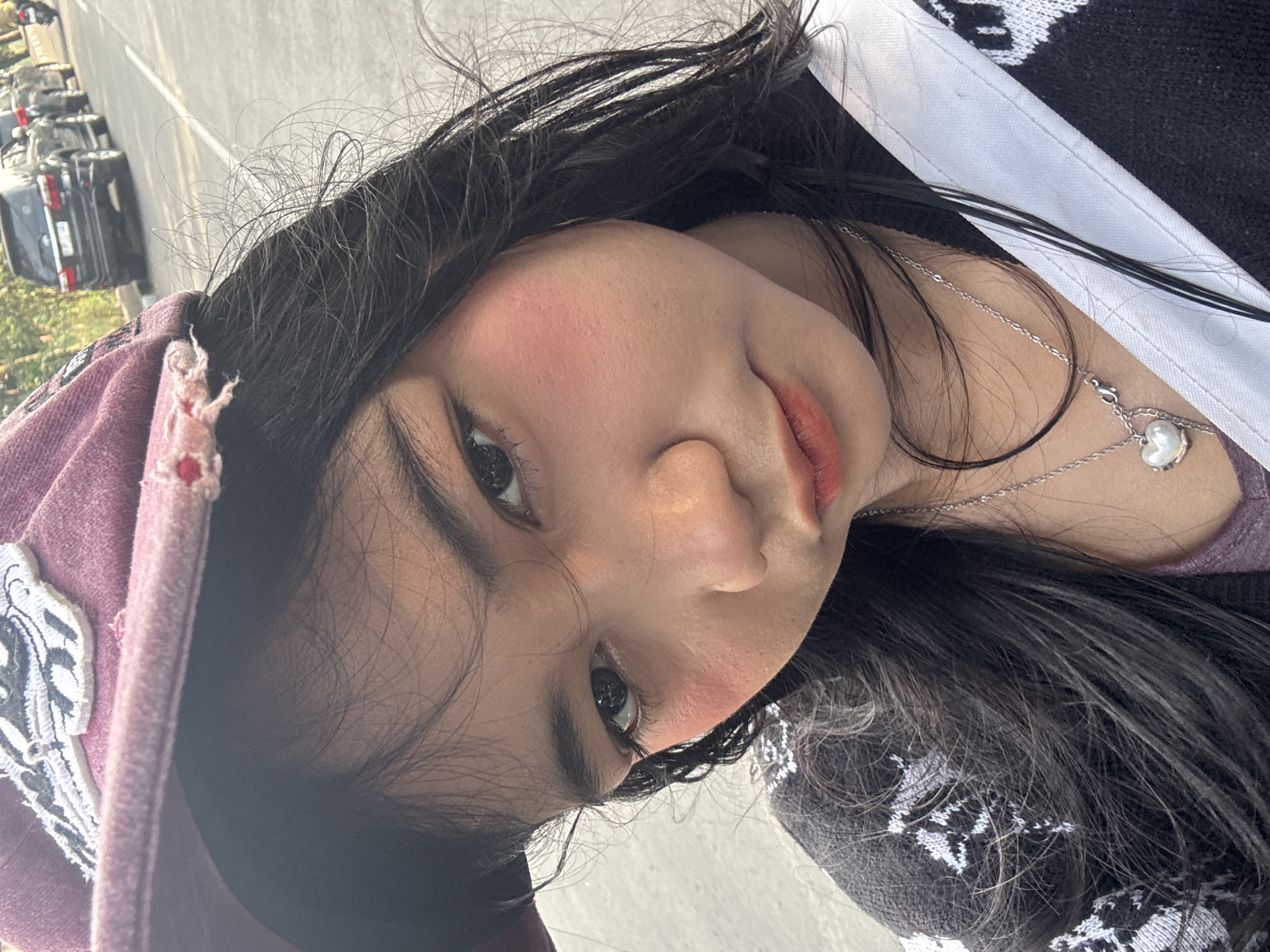 In North Jakarta, Indonesia, a young woman is captured in a close-up selfie, her face angled slightly towards the camera. She wears a maroon baseball cap with a black and white embroidered patch. Her dark hair, some strands appearing windswept, frames her face. She has dark eyes, prominent eyebrows, and rosy blush on her cheeks. Her lips are painted with a reddish-orange lipstick. She is wearing a black and white patterned cardigan over a purple top and a silver necklace with a heart-shaped pendant. The background shows a paved road with several parked cars and a distant motorcycle, suggesting an urban or roadside setting.