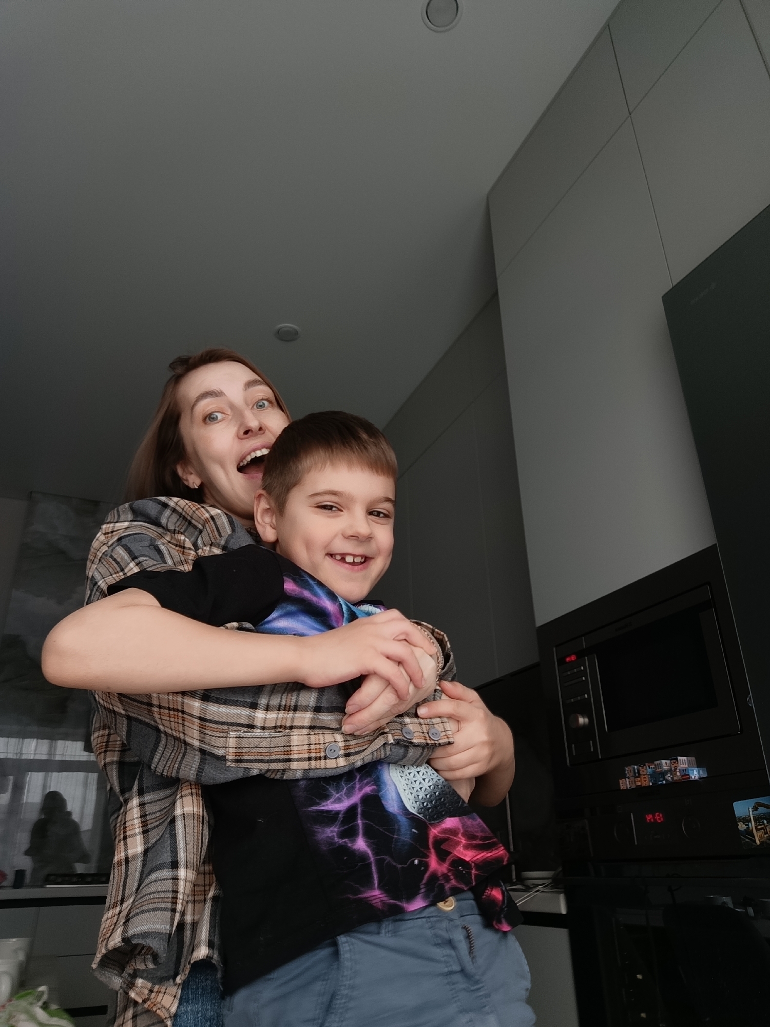 A woman and a boy, presumably a mother and son, are posing for a photo indoors. The woman has brown hair and is wearing a gray and brown plaid shirt. Her mouth is open in a wide smile, and her eyes are wide. She is holding the boy in her arms. The boy has short brown hair and is wearing a black shirt with a colorful graphic design. He is also smiling and has his arms around the woman. The location is a modern-looking kitchen. A built-in microwave is visible. The city of Chaikovsky, Russia, is not discernible from the image.