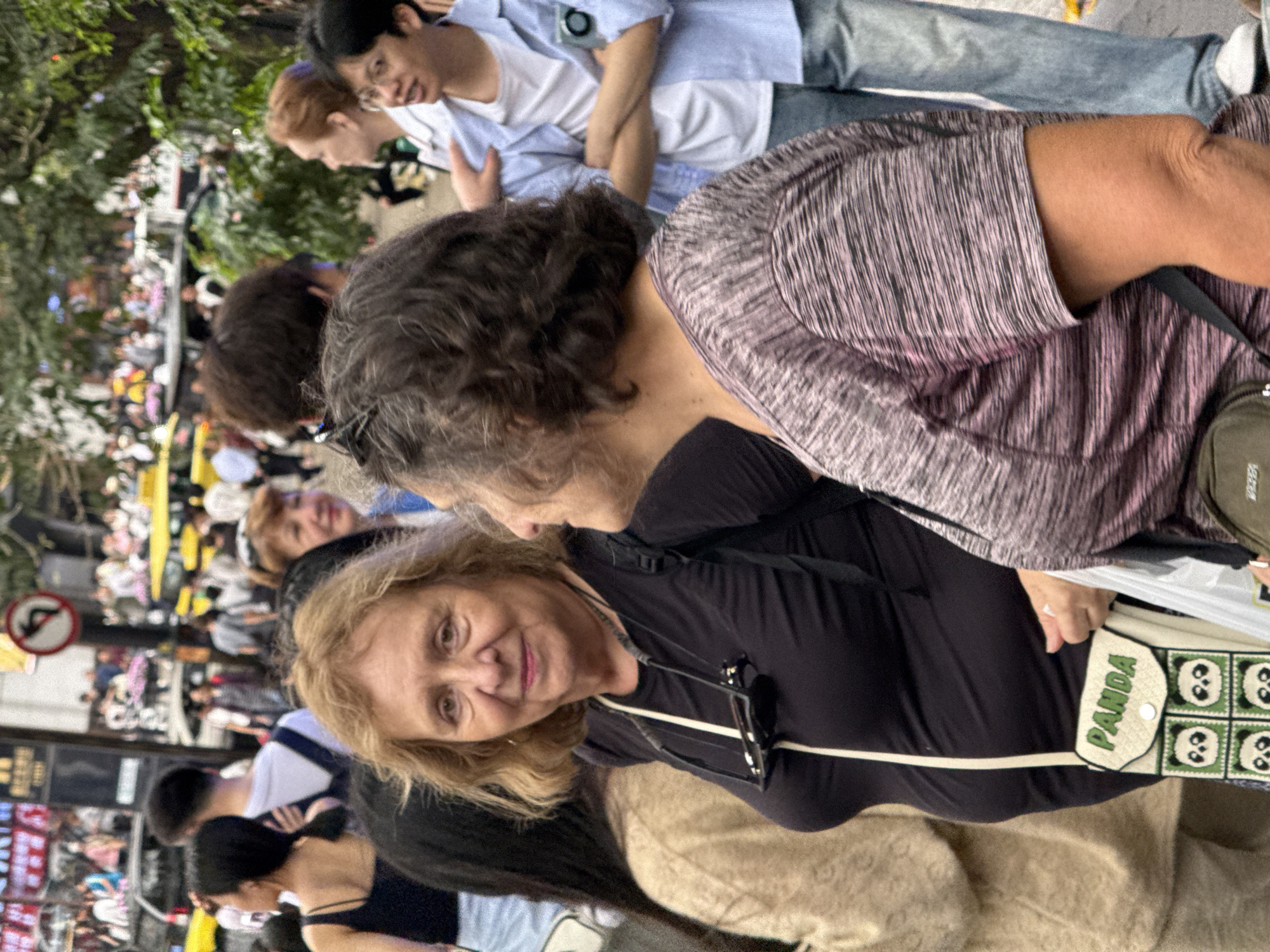 A crowded street scene, possibly in Yuzhong District, China, features multiple people. Dominating the foreground are two older women: one with blondish hair wearing a black top, the other with dark curly hair, wearing a striped top. The blondish-haired woman is looking at the camera with a slight smile. She has a beige bag with the word "PANDA" written on it, adorned with images of pandas. Behind them is a crowd of people in various poses, some looking at the camera, others engaged in conversation. A few Asian individuals are visible in the background. The setting is a bustling street with signs and possible storefronts along the sides.