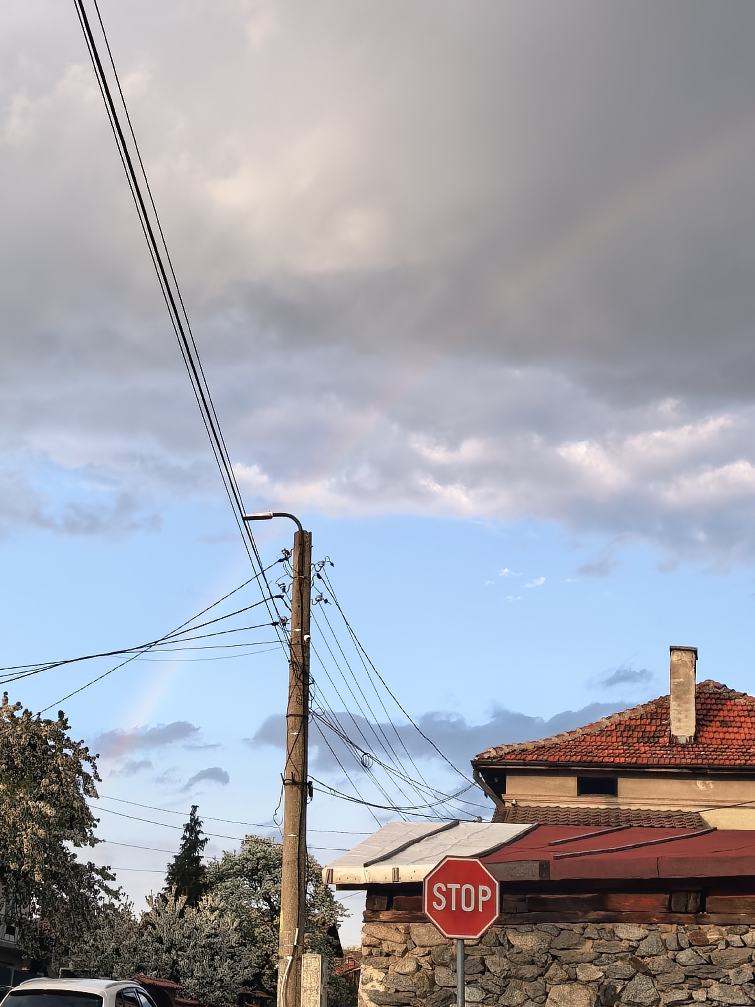 The image shows a residential scene under a partly cloudy sky with a faint rainbow visible. In the foreground, a red STOP sign stands near a stone wall, partially obscuring a building with red tile roofs. A wooden utility pole with wires spans across the image, and a street lamp is attached to the pole. Trees are visible to the left of the frame, and a portion of a white car is seen at the bottom left. In the background, another building with a red tile roof and a chimney is visible.
Location: Unknown, Unknown.