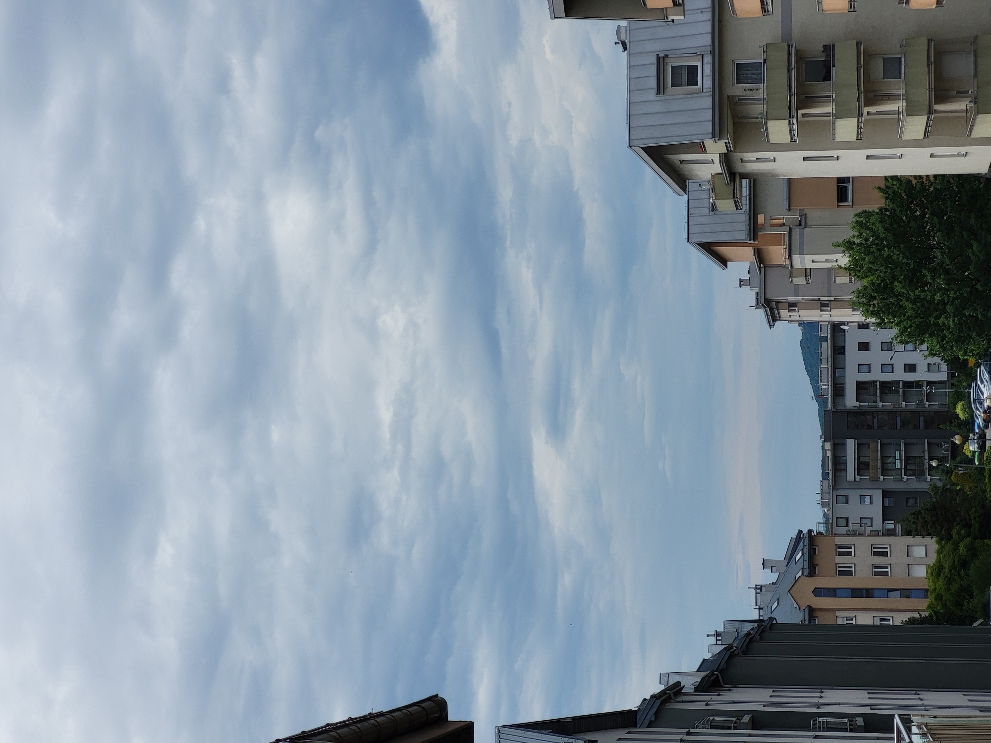 This is a vertical, outdoor, eye-level shot of a city skyline under a cloudy sky. The scene is dominated by apartment buildings with a variety of architectural styles and colors, including gray, beige, and dark gray. Some buildings feature balconies, while others have pitched metal roofs. Green trees and some parked cars are visible at the lower part of the image. The sky is filled with dense, gray clouds, creating a muted and overcast atmosphere.