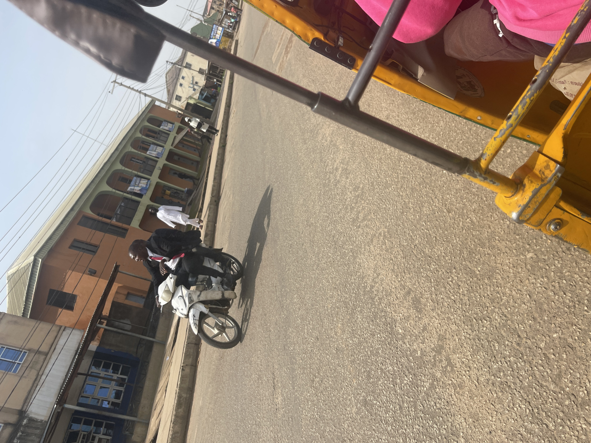 A man in a black suit and red tie rides a white motorcycle on a paved street in Rafin Par, Nigeria. He is looking to his right. In the background, there are buildings with orange and green facades. To the right of the motorcycle, a yellow and green auto-rickshaw is partially visible. The auto-rickshaw has a metal frame structure. A person wearing a pink garment is seated in the auto-rickshaw. The sky is clear and bright, suggesting it is daytime. The street is empty except for the motorcycle and the auto-rickshaw. The overall scene depicts a typical street view in an urban environment in Nigeria.