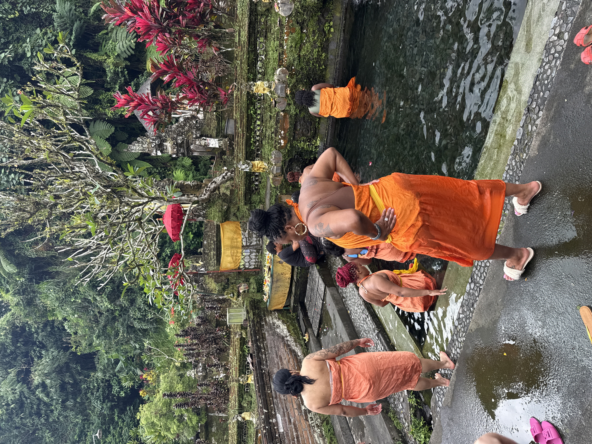 The image shows a group of people in what appears to be a Balinese bathing ritual, likely at the Tirta Empul Temple in Tegallalang, Indonesia. Several individuals, dressed in orange sarongs, are partially submerged in a pool of water. One person is standing at the edge of the pool with their hands on their hips. The scene is set in a lush, green environment, with tropical plants and traditional Balinese structures visible. The ground appears wet, suggesting recent rain or a damp climate.