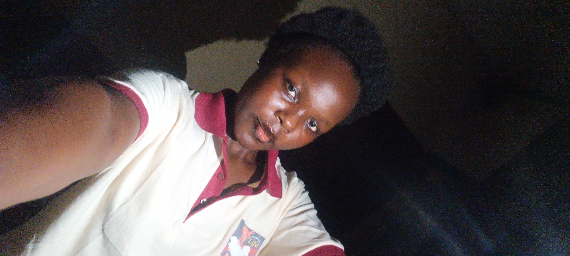 A young woman is taking a selfie in a dimly lit indoor location. The woman has dark skin and short, dark hair. She is wearing a tan short-sleeved shirt with a burgundy collar and trim. The shirt has an emblem on the lower front corner. The background appears to be dark, suggesting either a nighttime setting or an poorly lit room. There is not enough information to determine the location in the city of Onitsha, Nigeria.