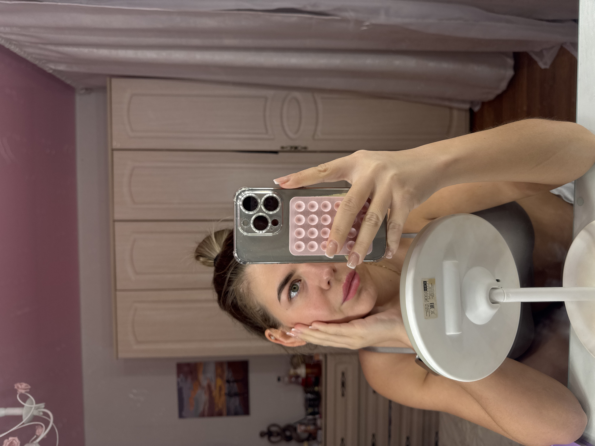 A young woman with fair skin, blue eyes, and blonde hair in a bun is taking a selfie in a mirror. She is holding a smartphone in her right hand, its pink case with circular patterns visible, and her left hand is resting on her cheek. The phone partially obscures her face, but her expression appears relaxed. Behind her is a wardrobe with paneled doors, a small table with decorative items, and a wall painted with light pink. The image was likely taken indoors, possibly in a bedroom. There is no information in the image to confirm a location in Chaikovsky, Russia.