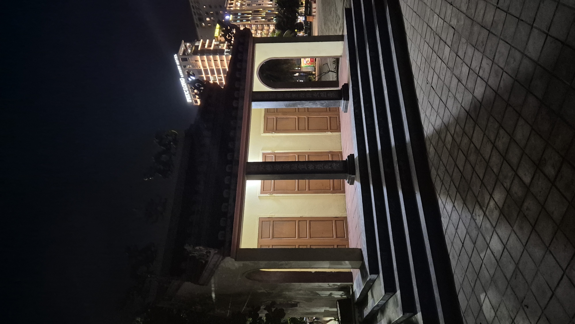 The image depicts a nighttime scene in Da Nang, Vietnam, with a traditional building featuring a dark, tiled roof supported by pillars. The building has several wooden doors and an open archway. A set of dark stone steps leads up to the building's entrance, and the ground in front is paved with square tiles that reflect light and cast shadows. In the background, a modern, lit-up building with the text "PHUC LONG LUXURY HOTEL" is visible, indicating an urban setting. The sky is dark and clear, suggesting it is night. There are no people clearly visible in the foreground, but there is a hint of movement or a person in the far left background near the building. The overall atmosphere is one of contrast between traditional architecture and modern city life.