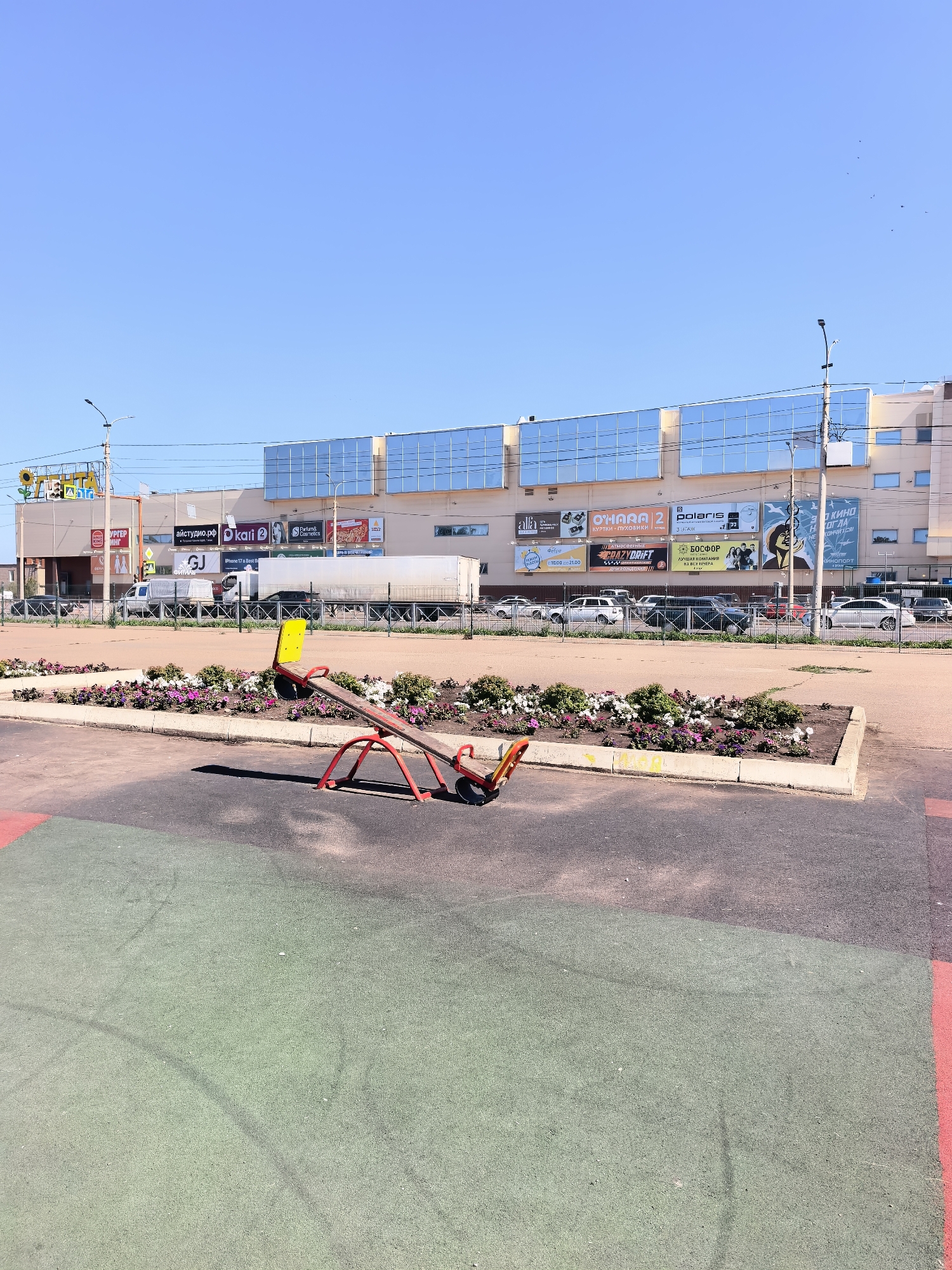A brightly lit daytime scene shows a red and yellow seesaw on a playground in Sterlitamak, Russia. The playground surface is composed of a green rubberized material with some dark patches and red accents along the edges. Beyond the playground, a concrete planter box filled with colorful flowers separates the play area from a paved walkway. In the background, a modern shopping mall with a facade of blue glass windows stands under a clear blue sky. Several vehicles, including cars and a white truck, are parked or driving in front of the mall. Power lines are visible overhead, strung between lampposts. The overall impression is a sunny day in a commercial area with a recreational space.