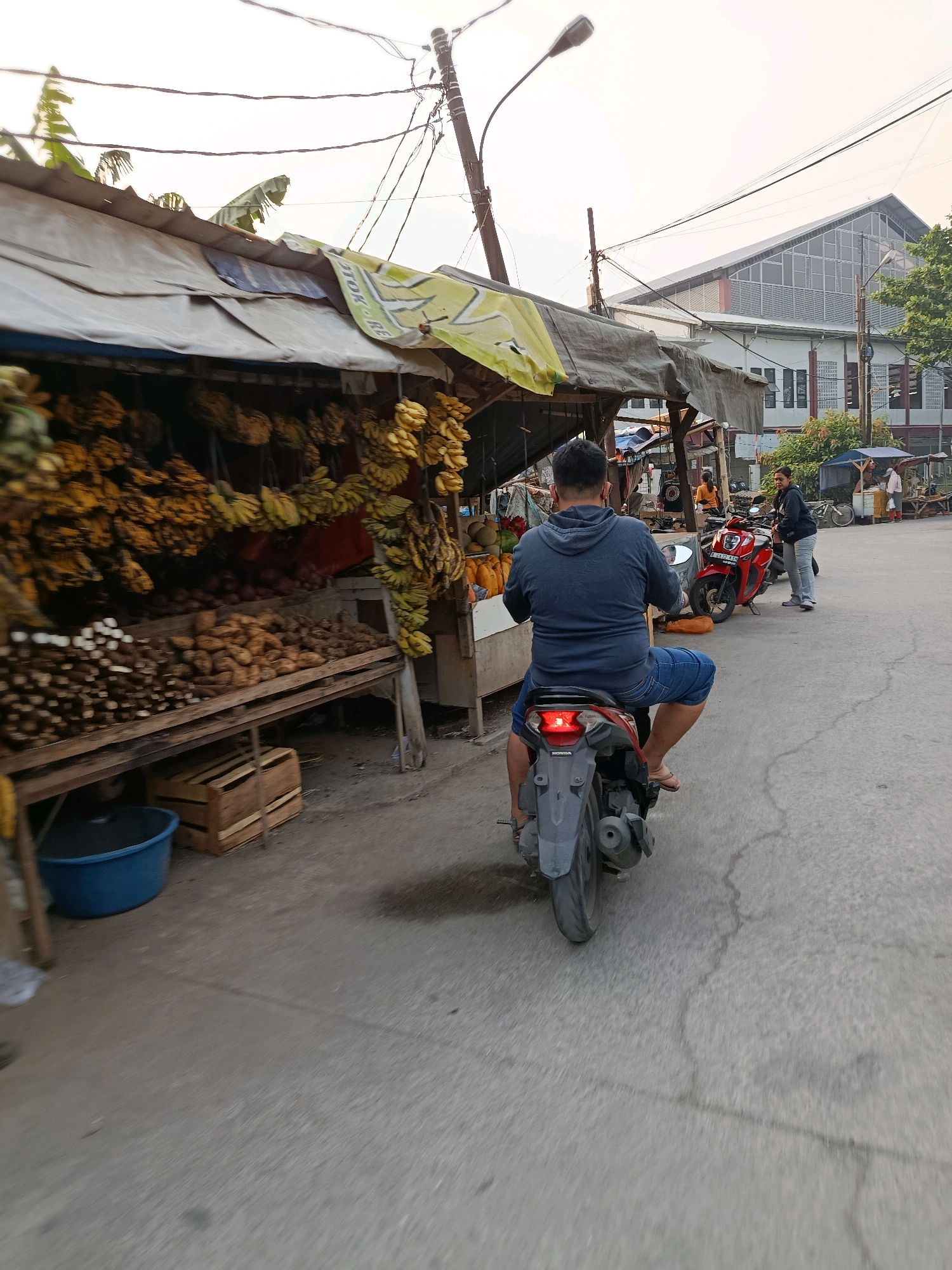 A man in a blue hooded sweatshirt and denim shorts rides a motorcycle away from the camera down a paved road lined with market stalls. The stalls are covered with tarps and display large quantities of bananas and other root vegetables. In the background, a modern building with a large, sloped, glass-paneled roof is visible. Other people are seen in the background, interacting at other stalls or with motorcycles. Power lines crisscross overhead. The sky is overcast. The scene appears to be a street market in the city of Bekasi, Indonesia.