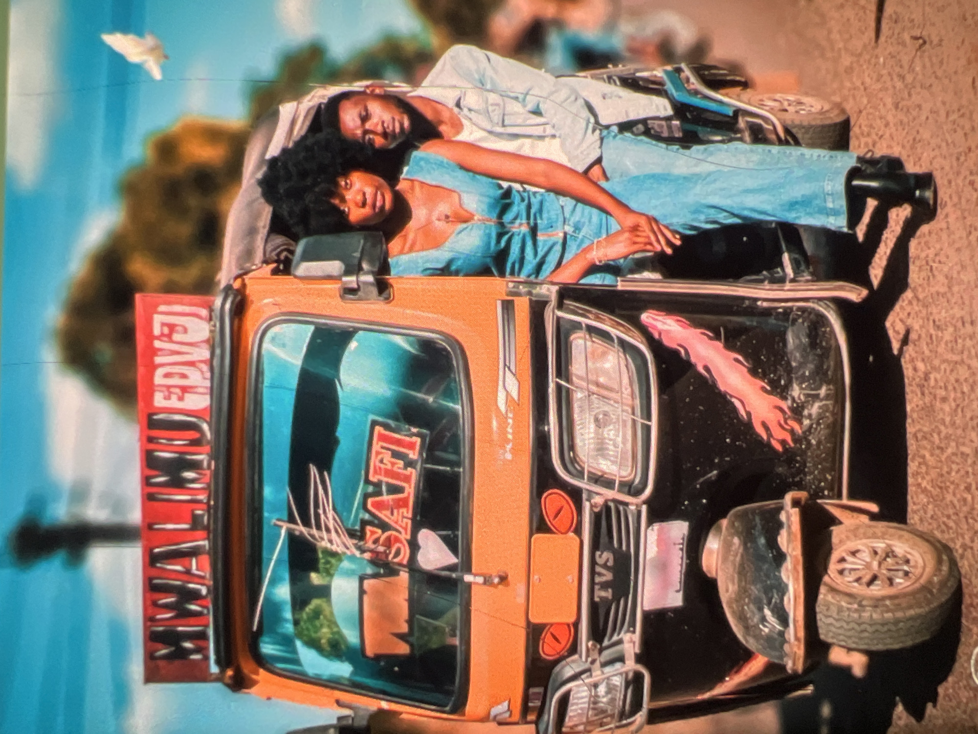 A young African couple is posed beside an orange and black auto-rickshaw, commonly known as a "Keke" in Lagos, Nigeria. The woman, wearing a denim jumpsuit, is seated inside the Keke, while the man, dressed in a light denim jacket over a white t-shirt, stands behind her, with his arm around her. The Keke has "MWALIMU FPVJ" written on its roof in red lettering, and "SAFI" is visible on the windshield. The vehicle's front features a "TVS" logo. The background shows a blurred urban environment with trees and a bright blue sky, with a white bird in flight. The overall image has a vibrant, possibly filtered, color palette.
