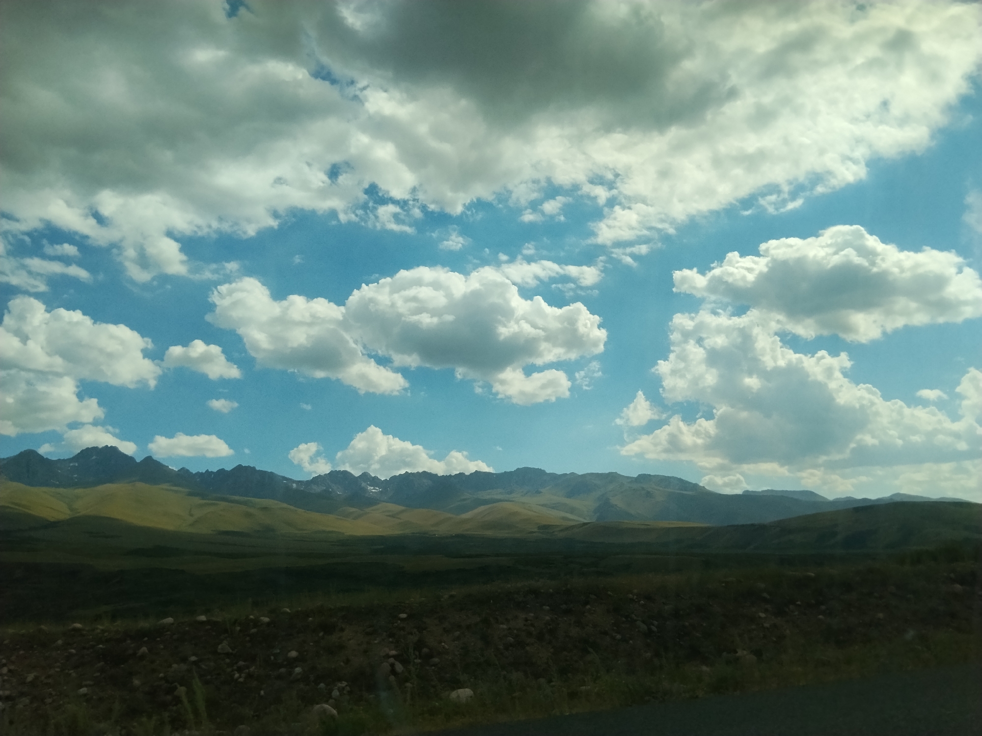 An outdoor, eye-level shot captures a vast, mountainous landscape under a partly cloudy sky. The foreground features a dark, rough terrain with scattered stones and sparse vegetation. Beyond this, rolling hills transition to a majestic mountain range in the distance. The sky is a vibrant blue, dotted with fluffy white and grey clouds. The scene appears to be in a rural, natural environment. The location is described as Unknown, Kyrgyzstan.