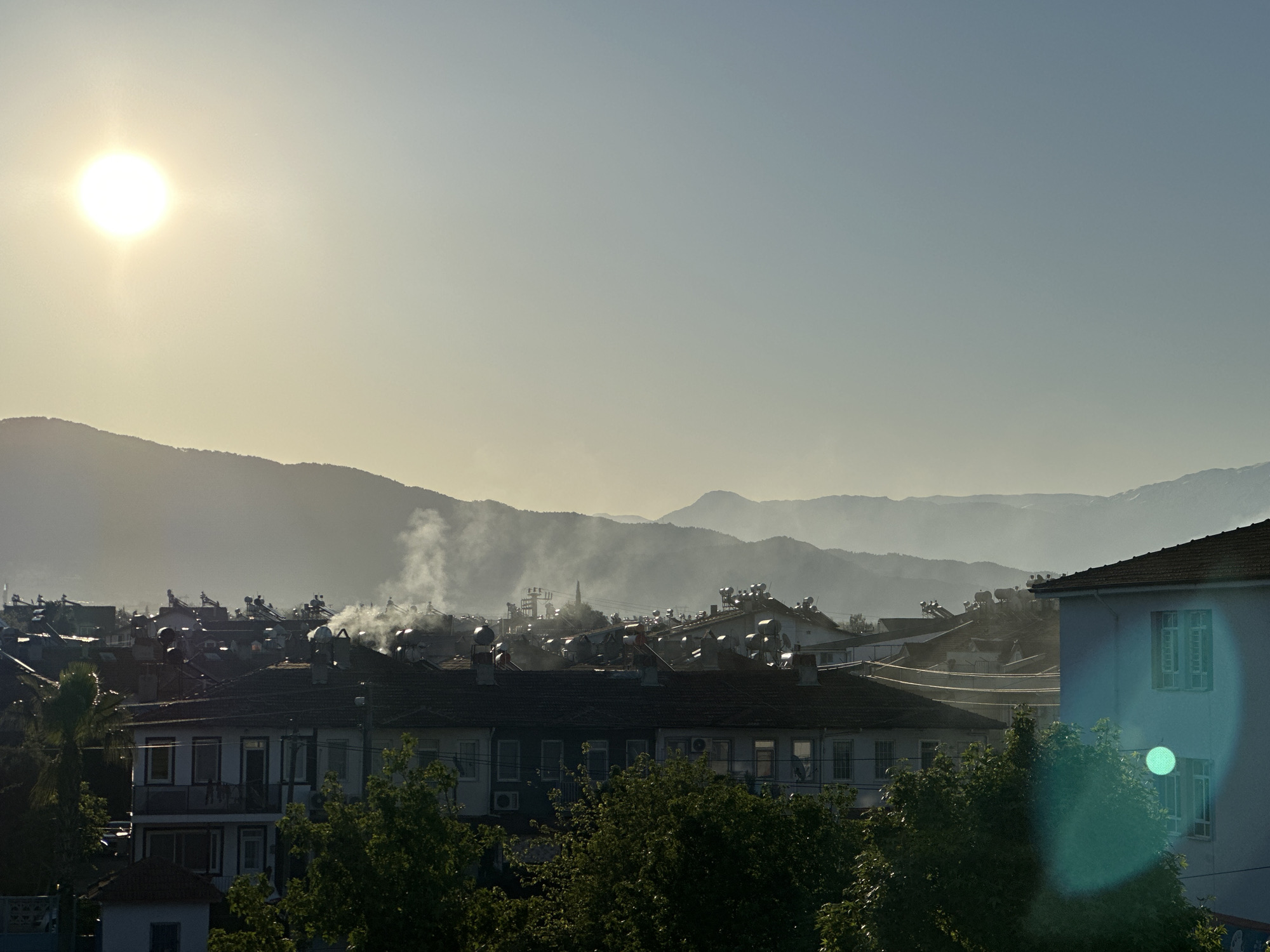 The image presents a wide, elevated view of a town or city, identified as Fethiye, Turkey, under a clear but hazy sky. The bright, intense sun is prominent in the upper left quadrant, casting warm light across the scene and causing a notable lens flare on the right side of the image.
In the background, a range of mountains appears hazy and layered, extending across the horizon. The mid-ground is dominated by a dense collection of buildings, primarily residential structures with reddish-brown tiled roofs. Numerous solar water heaters (cylindrical tanks) and some satellite dishes are visible on many rooftops, indicating local energy practices. Several distinct plumes of white smoke or steam are rising from various points among the buildings, suggesting activity such as heating, cooking, or burning.
The foreground features lush green trees that partially obscure some of the buildings. One light-colored building on the far right has visible windows and shutters, and it also shows a strong lens flare artifact. The overall impression is one of an active, populated area, likely captured during the early morning or late afternoon due to the low angle and warm tone of the sun. No people or visible text are discernible in the photograph.