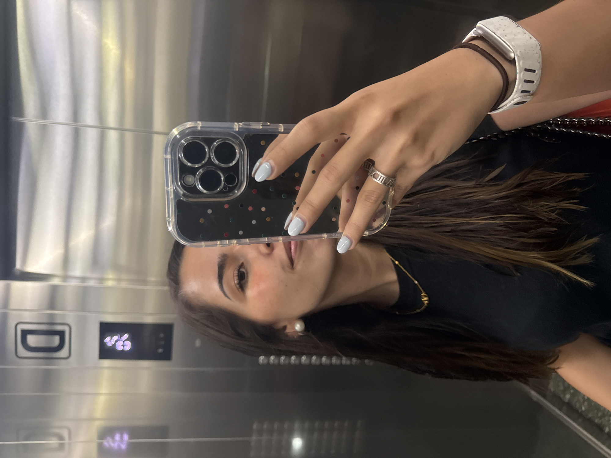 The image shows a woman taking a selfie in an elevator with mirrored walls. The elevator's control panel is visible in the reflection, indicating the floor number is "45". The woman is wearing a black t-shirt, a gold necklace, and a white smartwatch. She has light blue nail polish and a ring on her finger. The phone case is transparent with colorful polka dots and black camera lens frames. The location, San Bernardino Tlaxcalancingo, Mexico, cannot be verified as the photo could have been taken anywhere.