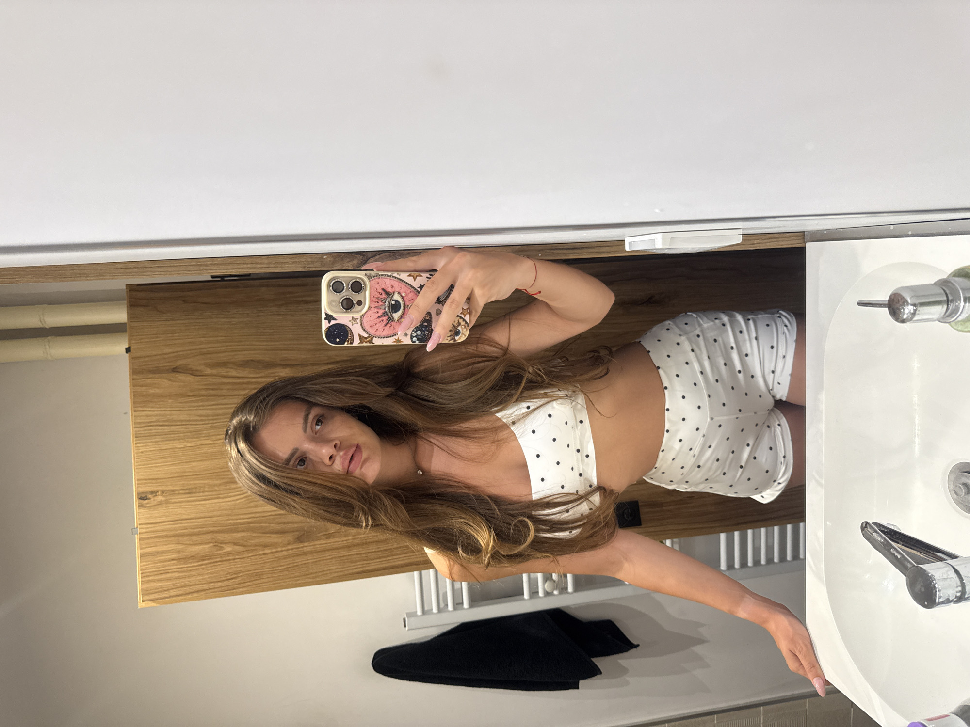 A young woman with long, brown hair is taking a mirror selfie in a bathroom. She is wearing a white polka dot bralette and matching shorts. The bathroom has a wooden vanity, a white sink with a chrome faucet, and a white towel rack with a black towel hanging on it. There are also some pipes visible on the wall behind her. The woman is holding a phone with a decorative case in her right hand, which is capturing the reflection. The scene appears to be in a modern residential bathroom. There is no indication of the location being in the city of Plovdiv, Bulgaria.