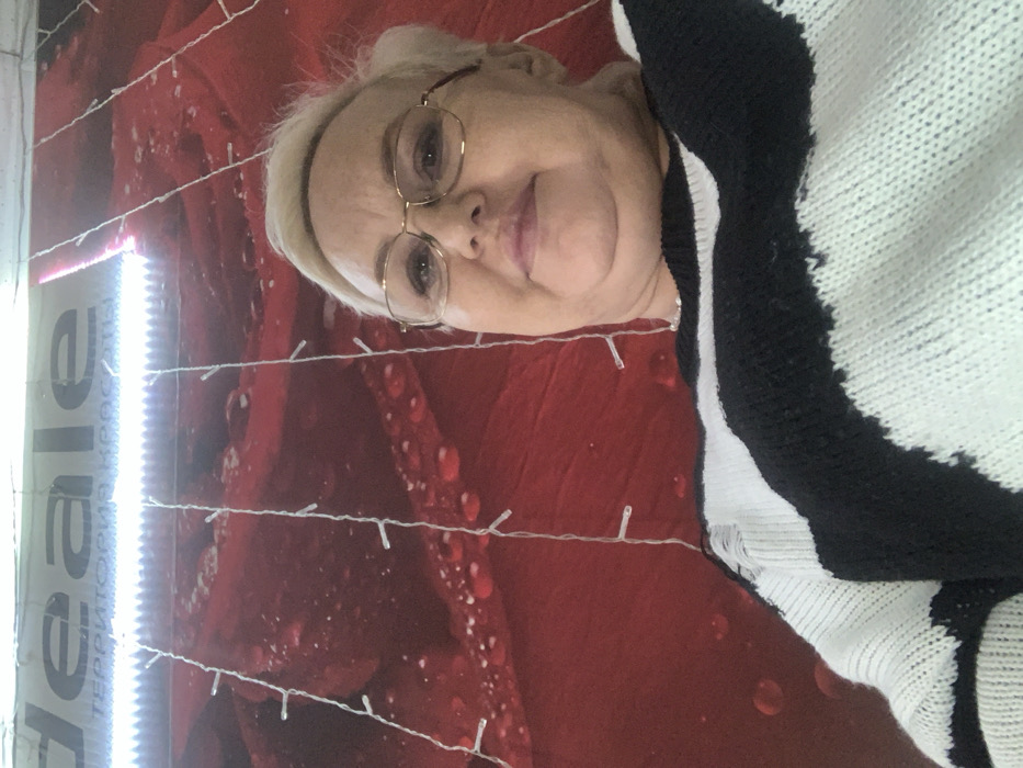 A blonde-haired woman wearing gold-framed glasses and a black and white striped knitted sweater, exhibiting a small tear on the left shoulder, is depicted from a low-angle, looking directly at the camera with a slight smile. The background consists of a red mural featuring rose petals with visible water droplets, overlaid with vertical strands of small white LED lights. Above the mural, a white sign displays "Ideale" in Latin script and "ТЕРРИТОРИЯ КРАСОТЫ" (Territory of Beauty) in Cyrillic, illuminated by a bright overhead light fixture. The scene is an indoor commercial setting, likely a beauty salon or similar establishment, located in the city of Chaikovsky, Russia.