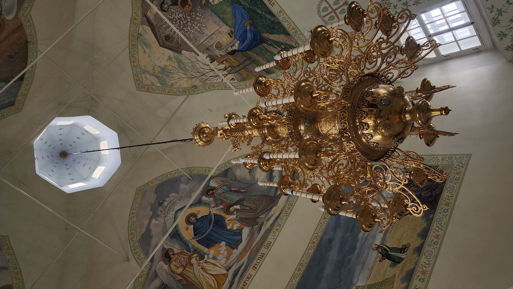 The image depicts the interior of what appears to be a church or religious building in Unknown, Unknown, with a focus on the ceiling and a large chandelier. The ceiling is adorned with religious murals or paintings. A central octagonal dome with windows lets in light. A gold-colored chandelier with multiple candle holders dominates the view, hanging from the ceiling. The overall environment suggests a place of worship with elaborate decoration.