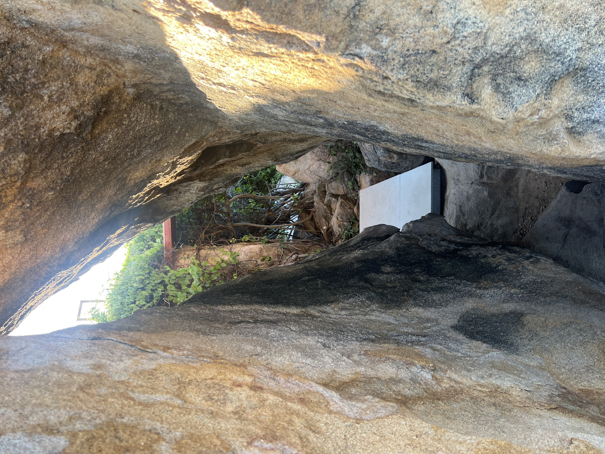 A pathway of light-colored concrete slabs leads through a narrow passage between large, weathered boulders. The rocks display a rough, textured surface with varying shades of brown, gray, and black. Lush green foliage, with vines clinging to branches, grows in the background, partially obscuring a distant structure with a roof. The scene is illuminated by natural light, casting shadows and highlighting the contours of the rocks. The location appears to be an outdoor environment, possibly a garden or a natural landscape feature within a developed area, in Vĩnh Hải, Vietnam.
