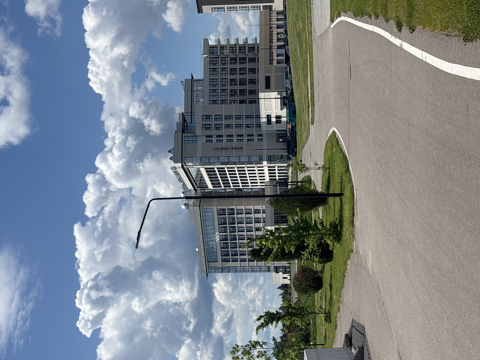 The image shows a street scene in Varaksino, Russia. A paved road curves from the foreground to the upper right, marked with a white line. To the left of the road is a green grassy area with small trees and bushes. A black lamppost stands on the edge of the grass. In the background, a modern multi-story building with glass windows is visible. The sky is blue with large, puffy white clouds. The image is oriented vertically.