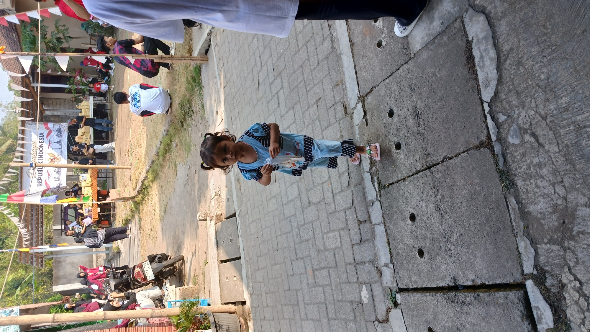 A young girl in a blue and black striped outfit stands on a paved walkway, holding a clear plastic bag. She has her hair in two pigtails and wears pink flip-flops. In the background, a street event is taking place. Several people are present, some are sitting, while others are standing near tables displaying goods. A motorcycle is parked on the left side of the image. A banner with "REPUBLIK INDONESIA" is visible in the background, suggesting the event might be related to Indonesian independence or a similar national celebration. The scene appears to be outdoors, with a natural light suggesting daytime. The paved walkway is made of interlocking bricks, and a concrete drain is visible in the foreground.