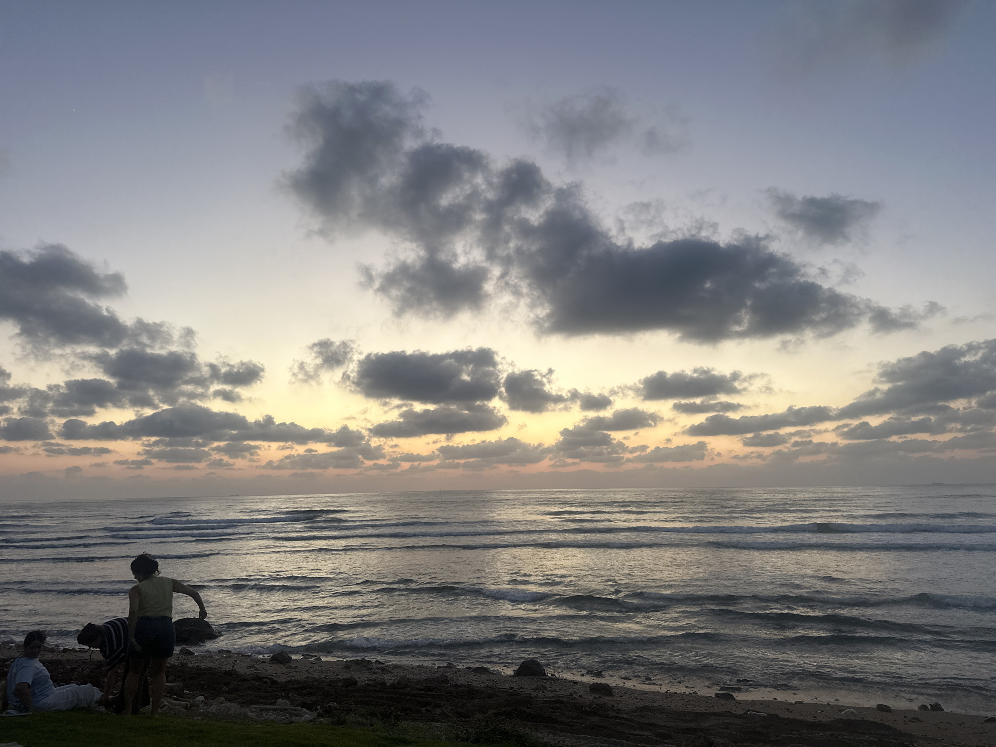 The image depicts a serene beach scene at dusk, likely in Haifa, Israel, given the context. The sky is a gradient of soft blues and yellows, transitioning from a pale lavender at the top to a warm, hazy orange near the horizon, indicating sunset or twilight. Scattered dark, fluffy clouds drift across the sky, adding texture and depth.
The sea is calm, with gentle waves rolling towards the shore, creating a repetitive pattern of white foam. The water reflects the muted colors of the sky. The shoreline is a mix of sand, rocks, and dark seaweed. In the foreground, on the beach and a grassy area, are three people. One person, wearing a light green top and dark shorts, is standing and facing away from the camera, seemingly interacting with something on the ground near the water's edge. Another person, wearing a striped shirt, is bending down next to them. A third person, wearing a white t-shirt, is seated on the grass to the left, looking towards the sea. Their posture suggests a moment of relaxation and contemplation, enjoying the natural beauty of the setting.
The overall mood is peaceful and tranquil, capturing the quiet beauty of a coastal evening. The absence of bright sunlight and the soft hues of the sky and water contribute to a sense of calm. No visible text is present in the image.