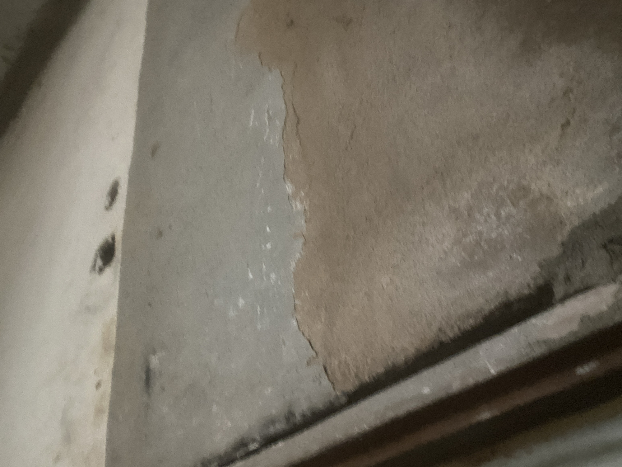 The image shows a close-up, low-angle view of a concrete surface that appears to be a floor or a wall. The surface is a light gray color and shows signs of wear and discoloration, with patches of reddish-brown staining and peeling in the lower right quadrant. To the left, a dark, vertical element, possibly a pipe or a structural support, runs along the edge of the concrete. In the upper portion of the image, the concrete transitions to a lighter, smoother surface, possibly a ceiling or a higher wall section, where several dark spots are visible. The lighting is dim, casting subtle shadows. The scene is devoid of any human presence or activity. This image depicts a section of deteriorated concrete infrastructure, potentially an interior surface within a building or a part of a public utility in Jauro Yinu, Nigeria.
