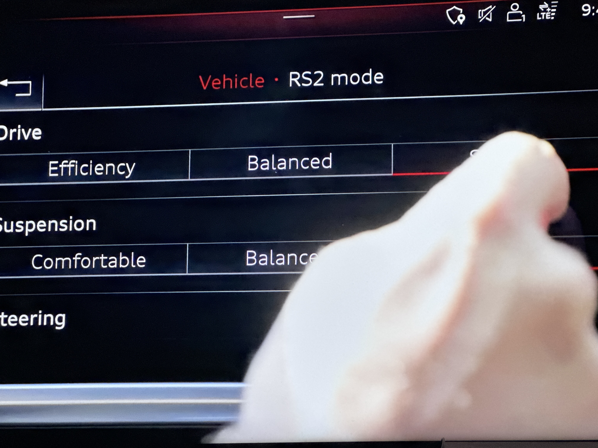 The image shows a car's infotainment screen displaying various driving modes. The "Vehicle - RS2 mode" is prominently displayed in red text. Below this, the screen shows categories such as "Drive," "Suspension," and "Steering." Under "Drive," the options "Efficiency" and "Balanced" are visible. Under "Suspension," "Comfortable" and "Balance" are visible. A blurred hand is interacting with the screen, touching the "Balanced" option for the suspension. The background is dark, consistent with a car's interior at night or in a dim environment. There is no visible indication of the city of Bezden, Bulgaria.