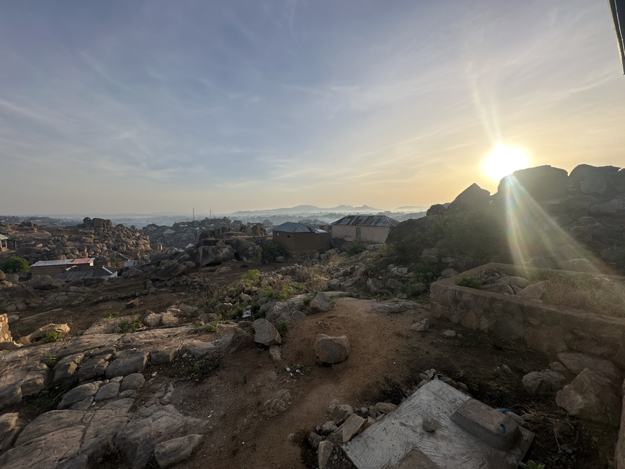 The image depicts a landscape in Jos, Nigeria, at sunrise. The foreground features rocky terrain with scattered boulders and sparse vegetation. In the midground, there are a few buildings with corrugated iron roofs, interspersed among more rocks and some greenery. In the background, a dense cityscape is visible, partially obscured by a light mist or haze. The sun is rising in the sky, casting a bright glare and creating silhouettes of the rocks in the foreground. A concrete structure is visible in the lower right corner. No people are visible in the image.
