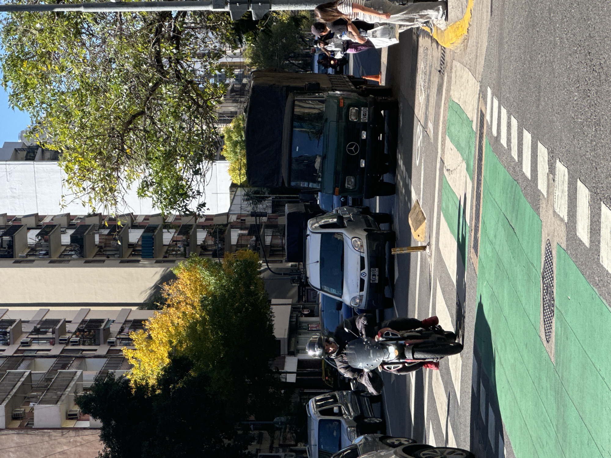 This image depicts a bustling street scene in Buenos Aires, Argentina, captured on a bright, sunny day. The view is rotated, showing the street, sidewalks, and buildings in an urban environment.
In the foreground, a person wearing a helmet and a dark jacket is riding a motorcycle on a paved road. The road features clear markings, including a distinctive green-painted bike lane to the right, bordered by white dashed lines, and a pedestrian crosswalk with white stripes. Further along the street, a dark green Mercedes-Benz truck, bearing the license plate "VZL 595," is visible, appearing to be stopped or parked. Behind the truck, a white Renault Kangoo van with the license plate "EFU 107" is also present, along with other vehicles partially obscured. An orange traffic cone stands near the truck.
On the right side of the street, two people are standing on the sidewalk. One individual, dressed in a striped shirt and jeans, is holding a white shopping bag. The street is flanked by multi-story buildings, characteristic of urban architecture, some featuring balconies. Trees with dense green and patches of yellow foliage add natural elements to the concrete landscape. The bright sunlight casts sharp shadows, indicating a clear sky and either mid-morning or early afternoon. The scene conveys the typical activity and rhythm of daily life in a vibrant city.
