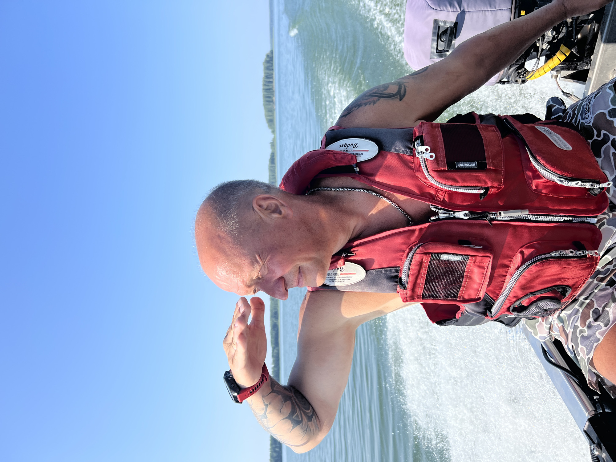 A bald man, wearing a red life vest and camouflage shorts, is on a boat on a sunny day. He is looking out at the water, shielding his eyes with his hand. A tattoo is visible on his left arm. The boat is moving, creating a wake behind it. In the background, there is a body of water and a distant shoreline with trees. The sky is clear blue. The image appears to have been taken in the village of Beregovoe, Russia.