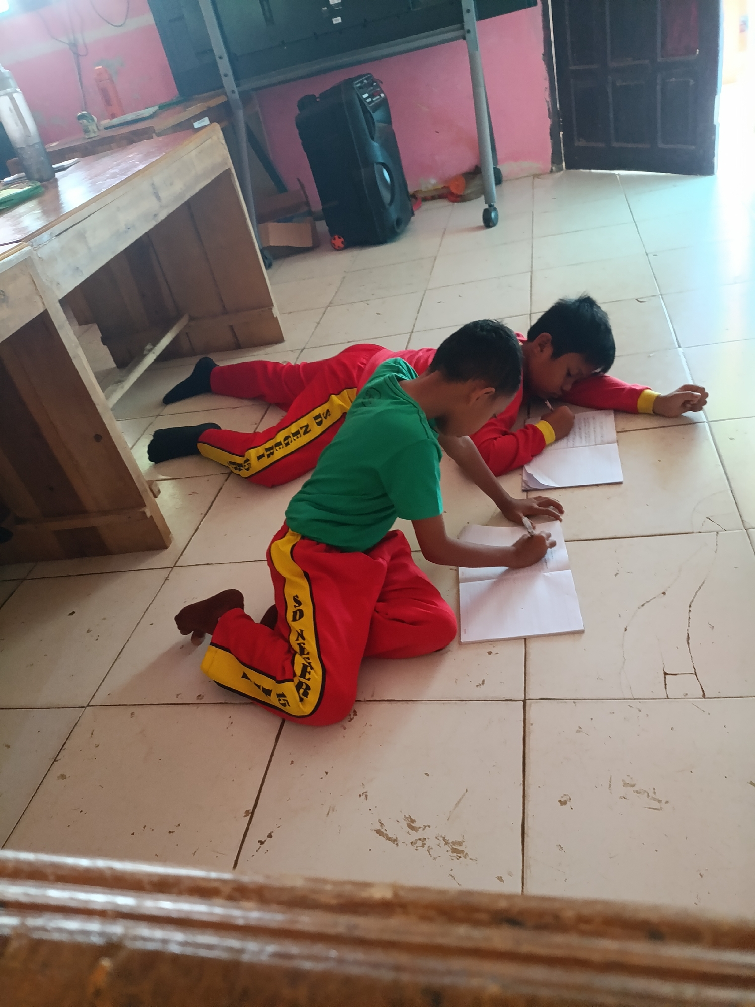 The image depicts two young boys in a classroom setting, likely in Sumberejo, Indonesia, engaged in writing activities on the floor. One boy, wearing a red uniform with yellow trim, is positioned on his knees and is actively writing in a notebook with a pen. He has dark hair and is wearing brown shoes. The second boy, also in a red uniform, is lying on his stomach next to him, with his head propped up on his arm, and is also writing in a notebook. He is wearing a green shirt and black socks. Both boys appear to be of elementary school age.
The environment suggests a typical Indonesian classroom. The floor is tiled with light-colored square tiles, some of which show signs of wear and tear. In the background, there is a wooden table with a slightly worn surface, and a large black speaker is visible. A tall, metal stand with wheels is also present, along with a pink wall. The lighting appears to be natural and somewhat diffused, indicating it might be daytime. The overall scene suggests an informal learning environment, possibly a break or a specific lesson format where students are encouraged to work on the floor. The uniforms, with "SD NEGERI" (State Elementary School) visible on the leg of one boy's trousers, confirm the educational context.