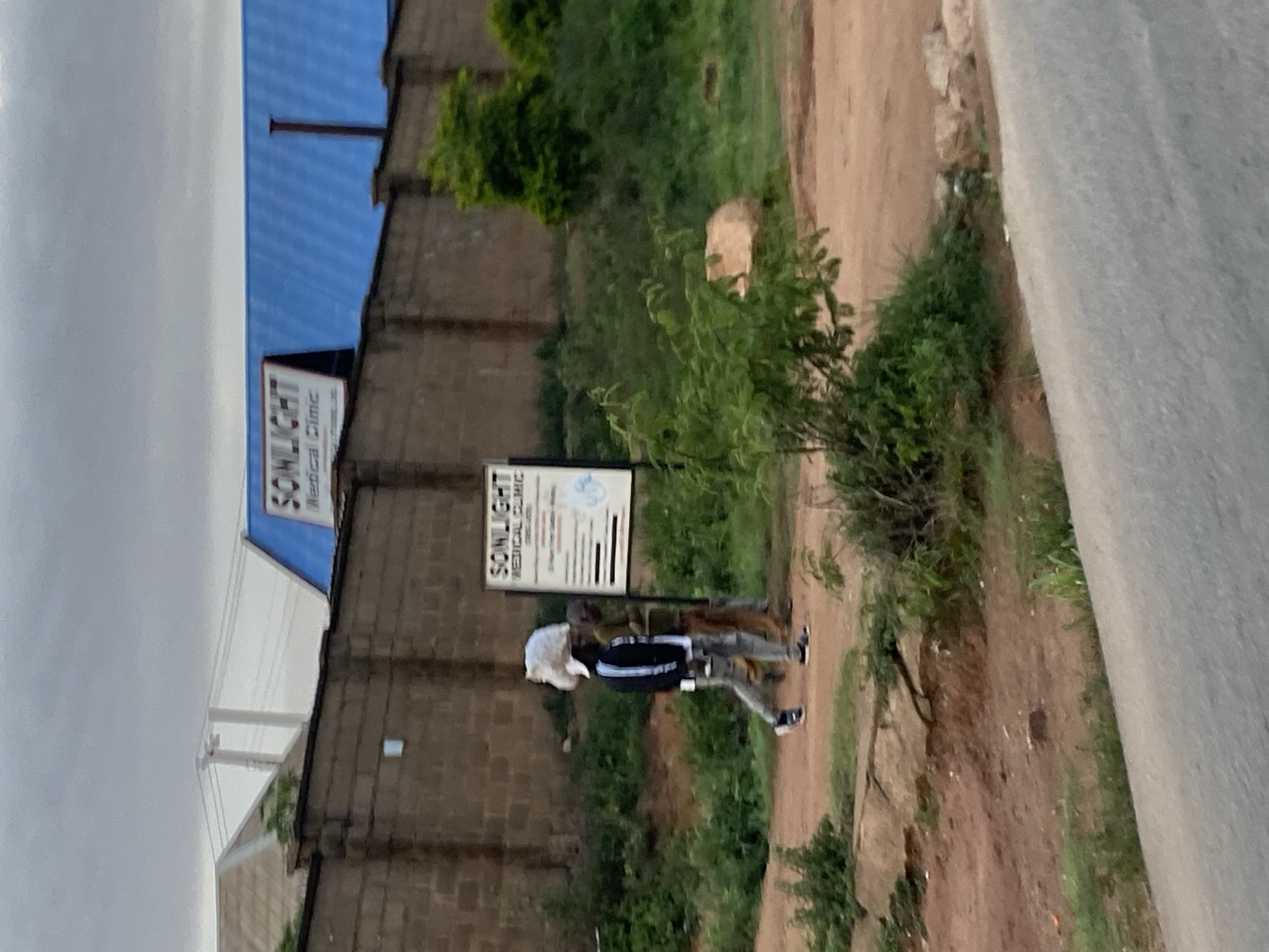 The image depicts a street scene in Jos, Nigeria, with a prominent brick wall along the side of the road. Two individuals are walking on the unpaved ground next to the wall. The person in the foreground, wearing a white cap, dark jacket, and light-colored pants, is carrying a large white bundle on their head. They are walking from left to right. Behind them, another person is walking in the same direction. A sign for "SUNLIGHT MEDICAL CLINIC" is visible on the wall. In the background, a building with a blue roof can be seen. The environment is dusty and somewhat overgrown with vegetation. The sky is overcast.