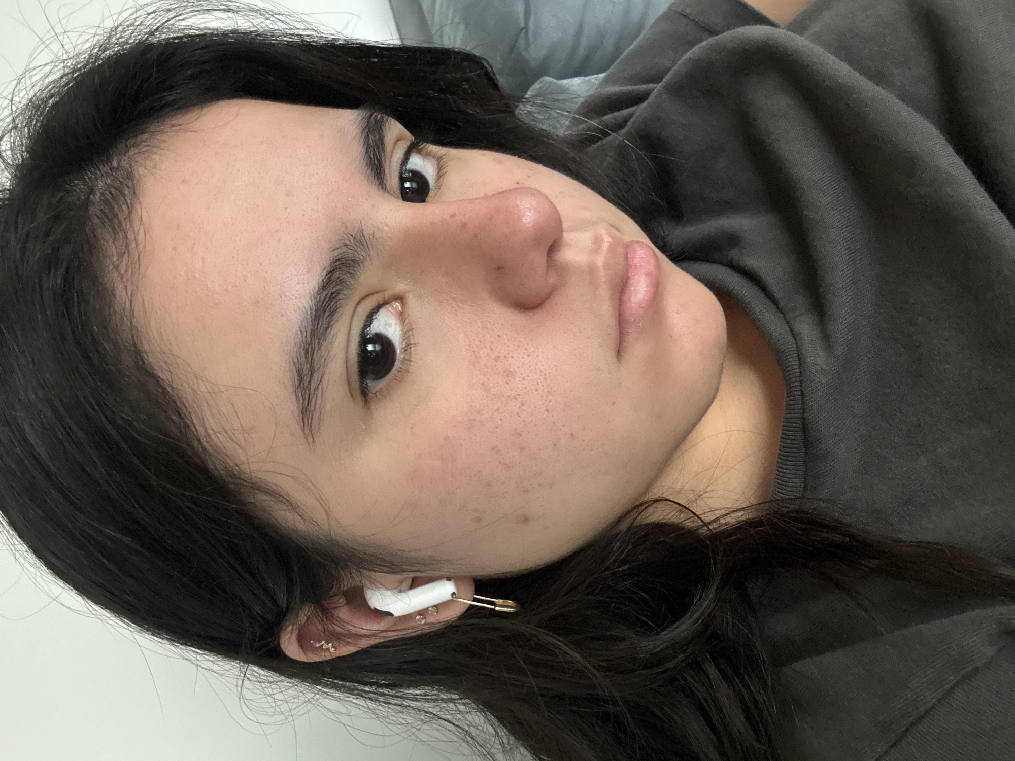 A close-up, diagonal portrait captures a young person, likely female, with dark, slightly wavy hair and dark eyes, resting indoors in what appears to be Austin, United States of America. She is looking directly at the camera with a neutral and calm expression. Her fair complexion shows visible pores and minor redness or blemishes on her cheeks and nose. Her lips are a natural light pink, slightly parted.
She is wearing a simple dark grey t-shirt or sweatshirt, the ribbed neckline of which is visible. A prominent detail is a white earbud positioned in her left ear, uniquely secured with a gold-colored safety pin. Her left ear also features multiple small, delicate piercings arranged along the lobe and helix. The background consists of soft, light and dark grey fabrics, suggesting she is resting on a bed or sofa. The indoor lighting does not indicate a specific time of day or weather. There is no visible text in the image.