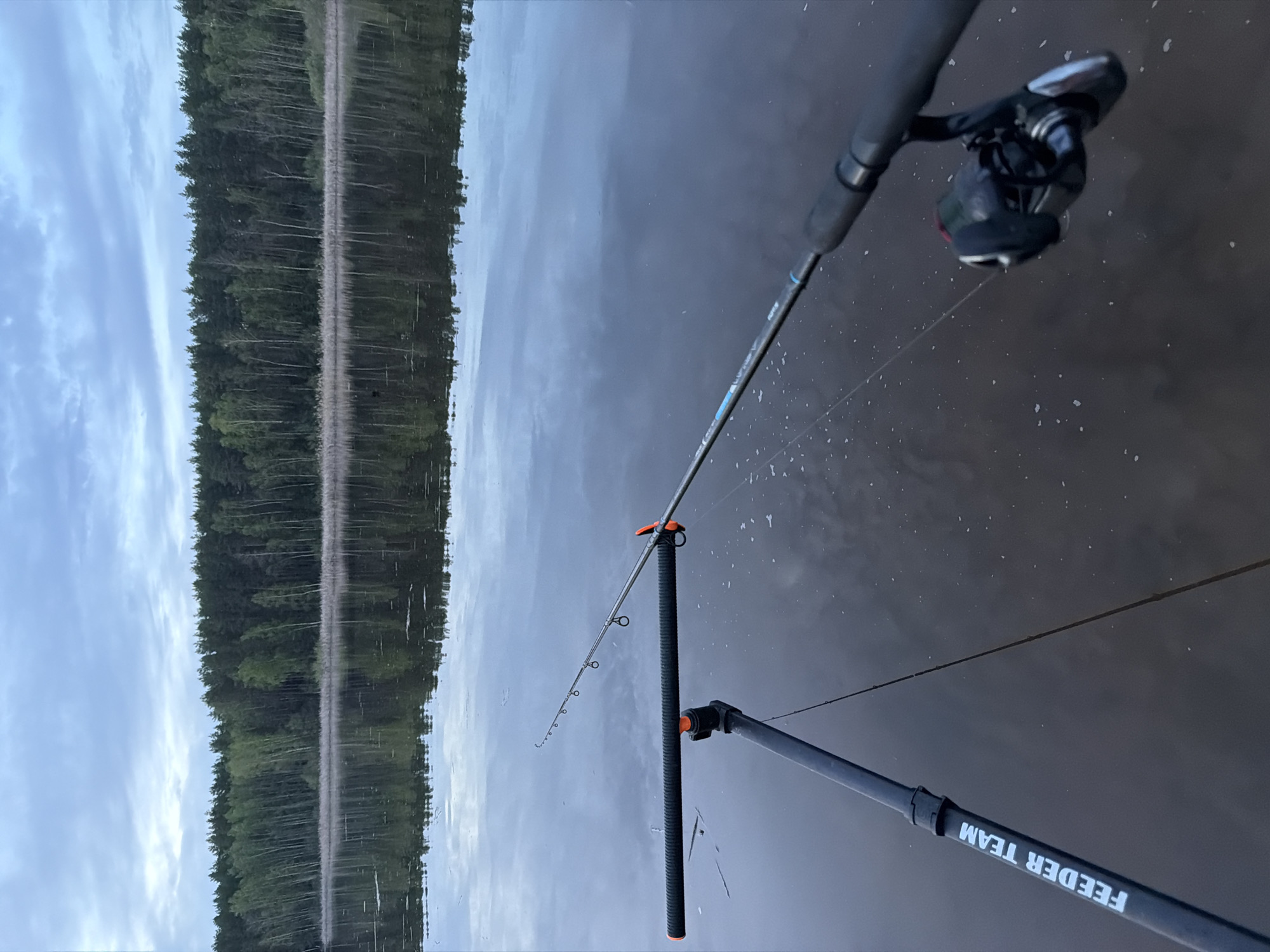 The image depicts a fishing scene, likely at a lake or river, in a natural environment. A fishing rod is positioned in the foreground, supported by a feeder team stand. The rod has a reel attached, and the line extends into the water. In the background, trees are reflected on the water's surface. The sky is visible, with reflections in the water as well. The presence of a "FEEDER TEAM" stand suggests a fishing competition or team activity. While the location of the image is possibly in the city of Chaikovsky, Russia (as requested), there is no specific visual information to confirm the location based on the image provided.