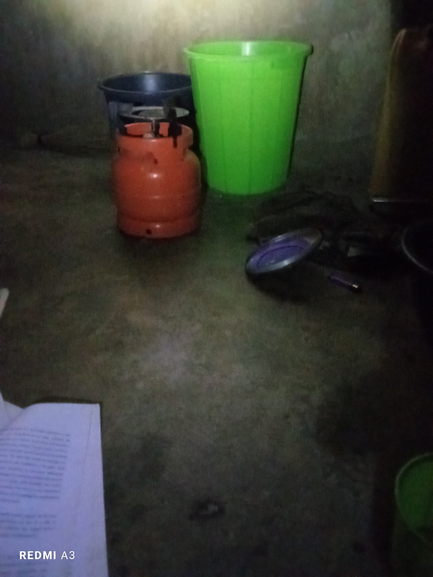 An orange gas cylinder is placed on the floor next to a black bucket and a green bucket. To the right of the gas cylinder, a small cooking stove is visible. In the foreground, a partially opened book or paper lies on the floor, with the text "REDMI A3" printed at the bottom left corner of the image. The scene appears to be indoors, possibly a storage area or a kitchen, with a rough, textured wall in the background. The lighting is dim, suggesting it might be nighttime or the room is poorly lit.