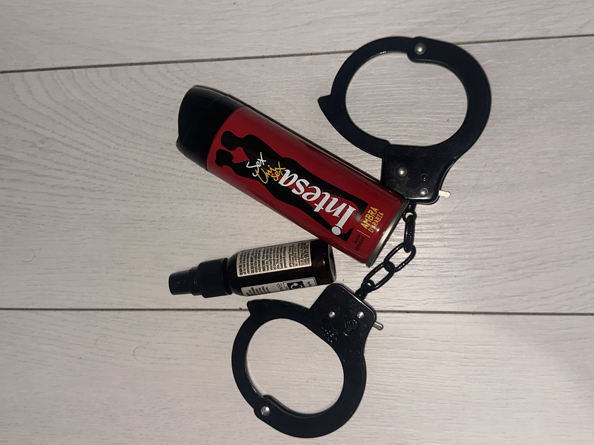 The image displays a still life arrangement of three objects on a light-colored wooden floor. The floor consists of planks oriented vertically, with visible wood grain and a prominent seam running through the center of the frame.
The objects present are:
1. One pair of black metal handcuffs, linked by a chain. The handcuffs are positioned with one cuff to the upper left and the other to the lower right, with the chain extending between them. Both cuffs appear to be open or partially open.
2. One small, dark brown or black glass spray bottle with a black pump dispenser. The bottle features a white label with text, partially legible as "BEARD OIL" and "Ambra," indicating it contains a product for personal care.
3. One red aerosol can with a black cap. The can displays "Intesa" in large white lettering, "Sex Unisex" in smaller text above it, and "AMBRA ARABIA" near the bottom. The design also features black silhouettes of a man and a woman.
The spray bottle is situated to the upper left of the red aerosol can. The red aerosol can is oriented diagonally, with its top pointed towards the upper left. The chain of the handcuffs passes underneath the bottom portion of the red can and is adjacent to the spray bottle. No people or discernible actions are depicted. The objects are stationary. The precise location in the city of Sofia, Bulgaria, cannot be determined from the visual information provided in the image.