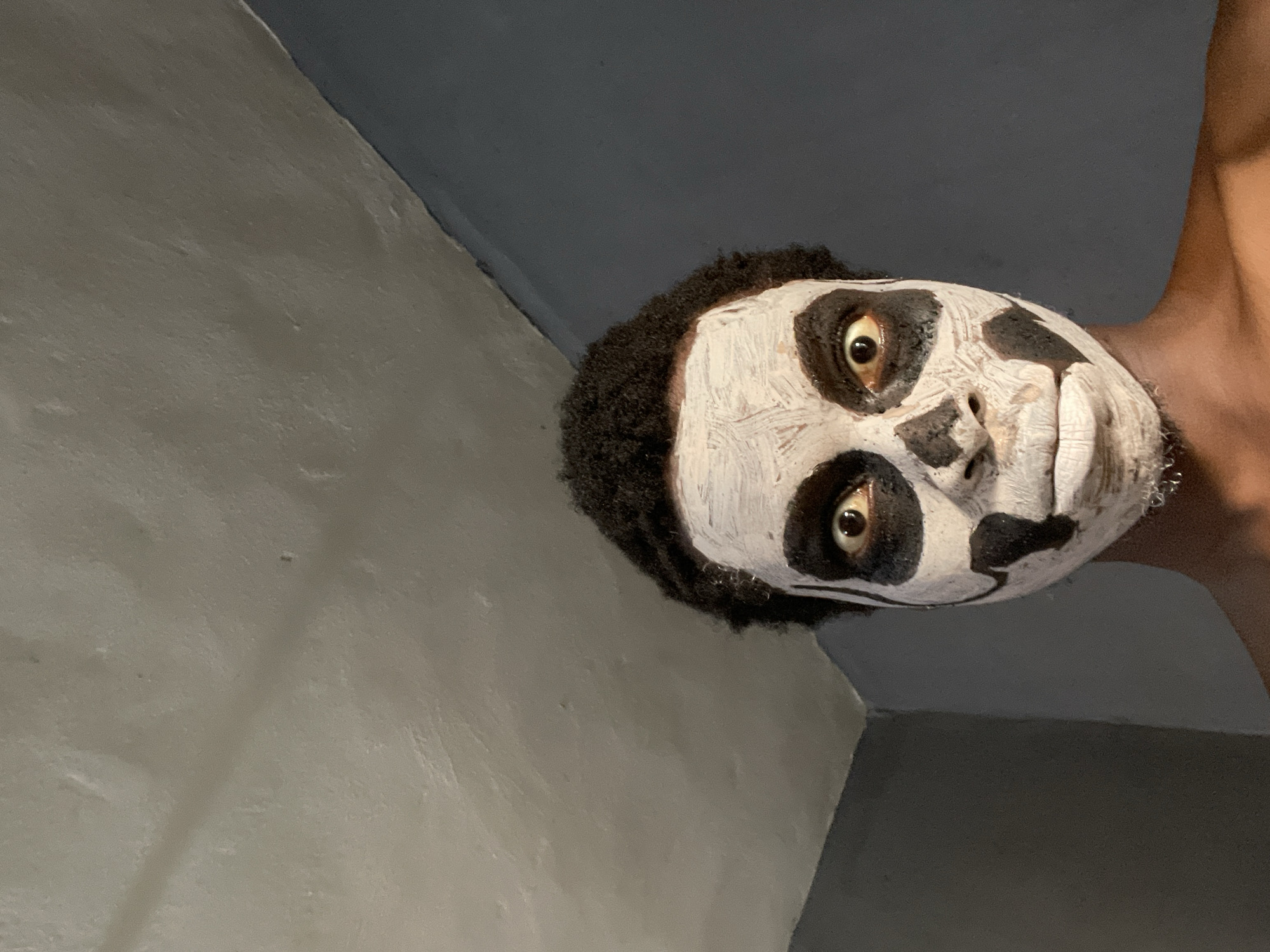 A close-up shot from a low angle shows a person with dark skin wearing white face paint with black accents, resembling a skull. The person has short, dark, curly hair. The background is composed of light grey and dark grey walls, implying an indoor space. There is no indication of location or other context, making it impossible to state that this image was taken in Onitsha, Nigeria.