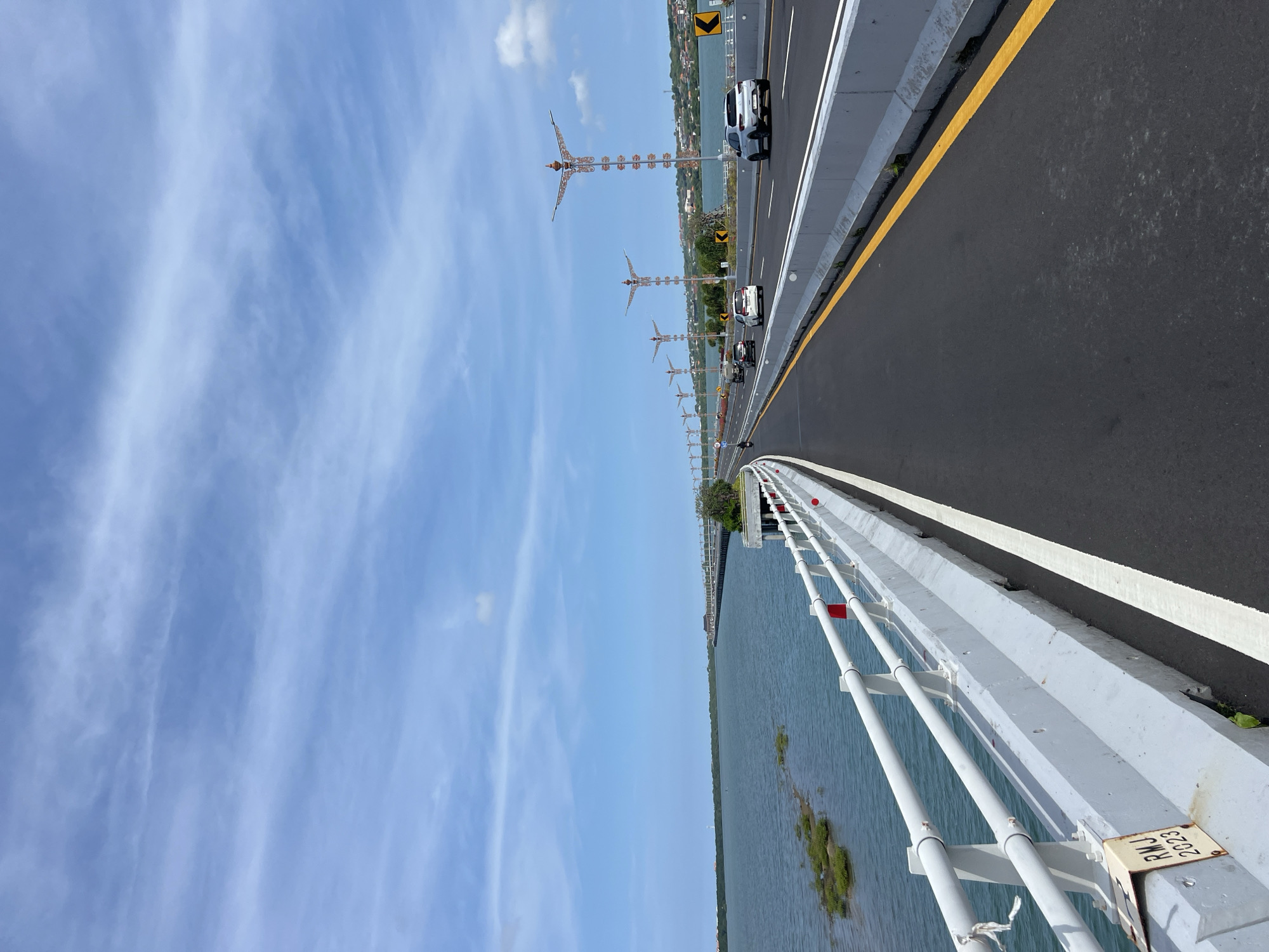 A daytime shot from an elevated position on a bridge with a view of the sky and water. On the right side of the image is the bridge roadway, painted black with a yellow line on the right edge and white stripes indicating lanes. There are cars driving along the roadway. On the left side, there is a white painted metal barrier with red reflectors, separating the road from the water. Below the barrier is a body of water. In the distance, there are decorative street lamps that stand along the roadway. The sky is blue with wispy clouds. The bridge is in Nusa Dua, Indonesia.