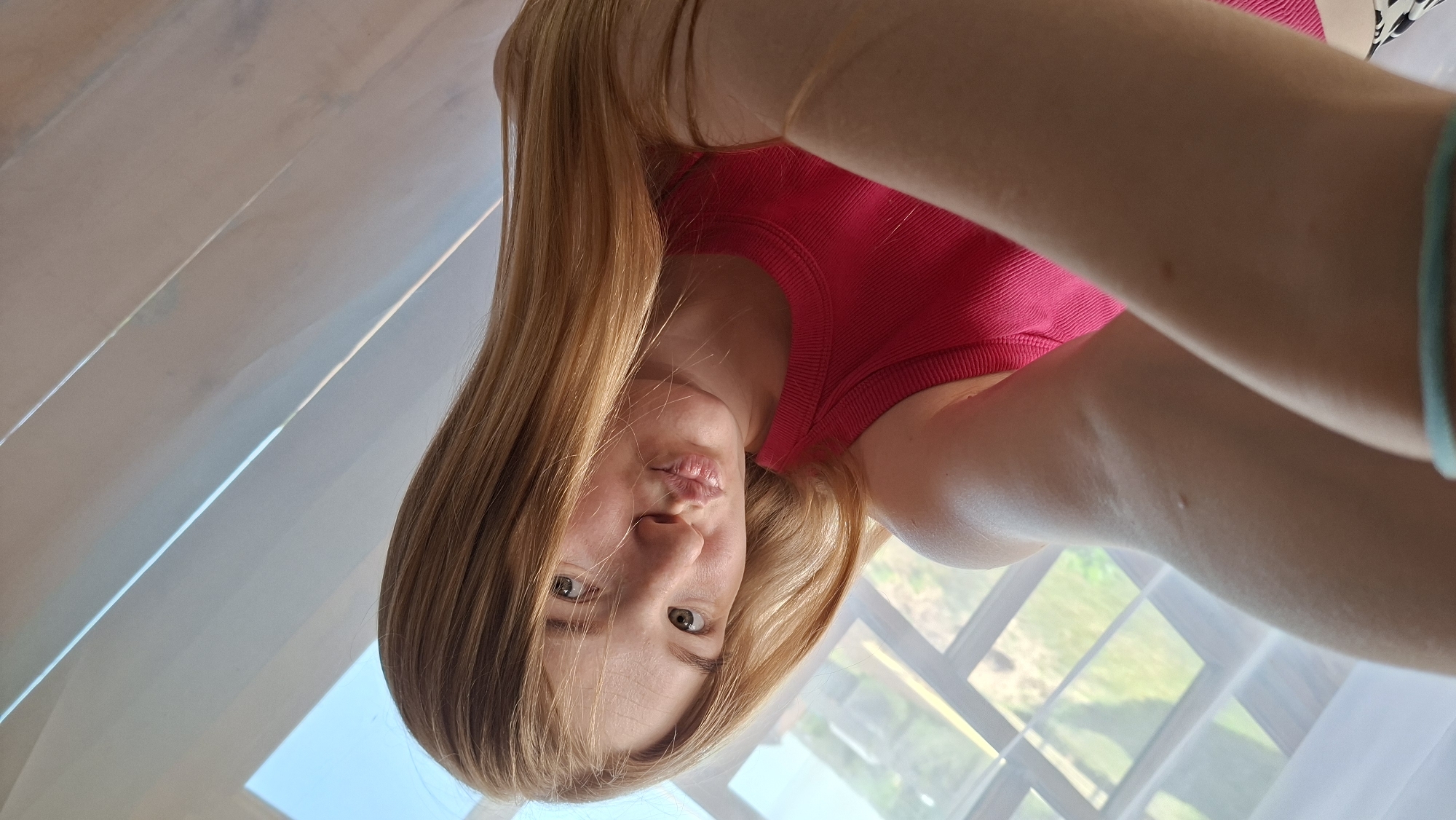 The image depicts a person with long, light brown hair wearing a pink top, positioned in front of a window. Outside the window, greenery is visible. The person appears to be indoors, possibly in a room with a light-colored wall or curtain. The location, Большой Букор, Russia, cannot be verified from the image.