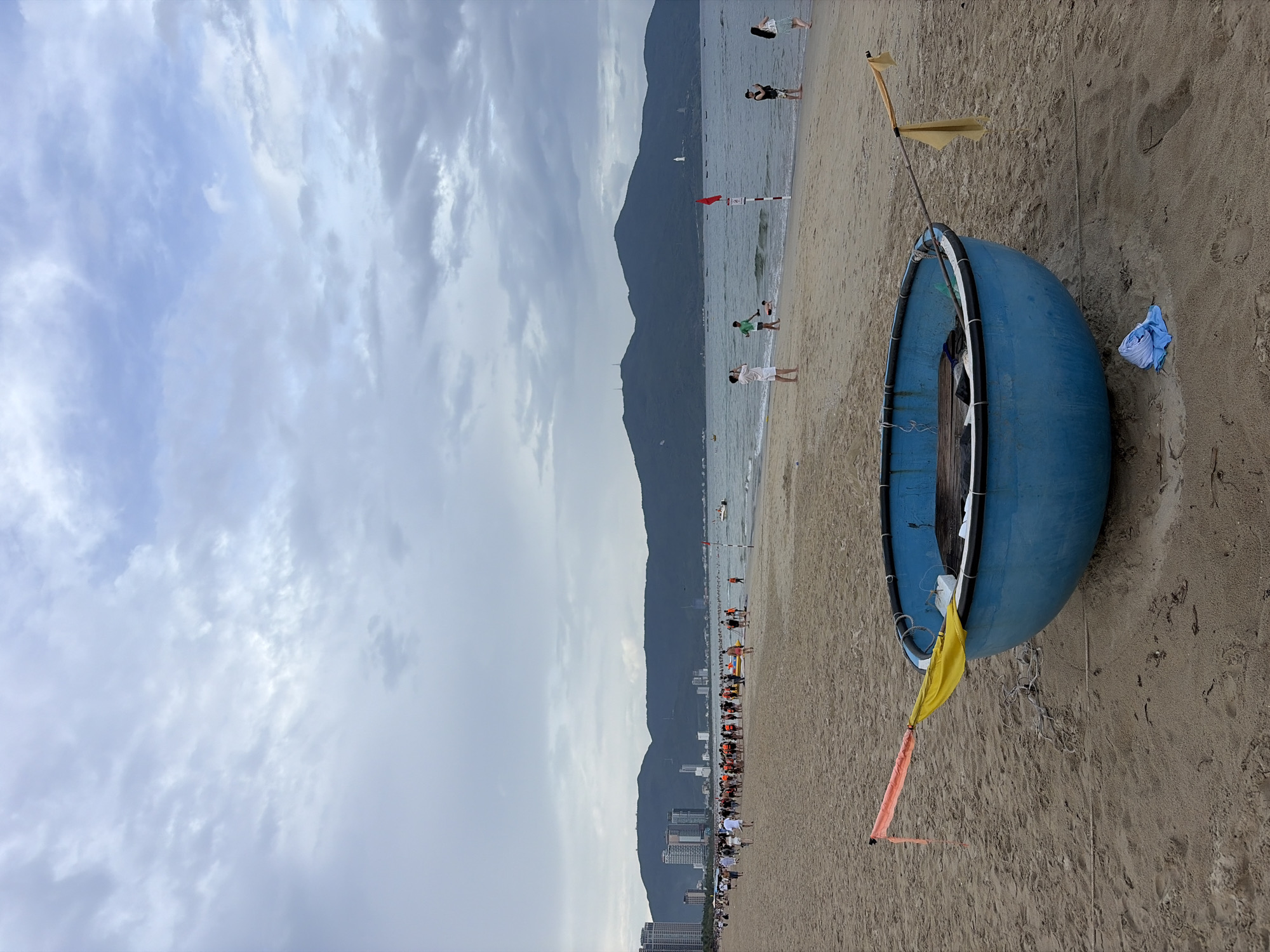 A wide shot captures a tranquil beach scene in Đà Nẵng, Vietnam. The sky is a canvas of overcast clouds, with a gentle, hazy light illuminating the landscape. In the foreground, a striking blue, round boat rests on the sandy shore, adorned with a vibrant orange and yellow flag. A few scattered items, including a folded blue cloth, lie on the sand near the boat.
Further back, the beach stretches out, populated by a diverse group of people. Some are engaged in activities near the water's edge, while others stand in groups along the shoreline. The sea, a calm expanse of blue, meets the horizon where distant, lush green mountains rise, their peaks softened by the atmospheric haze. Along the coastline, a city skyline emerges, marked by modern buildings. The overall atmosphere is one of peaceful coexistence between nature and urban development, with a hint of community activity on the beach.