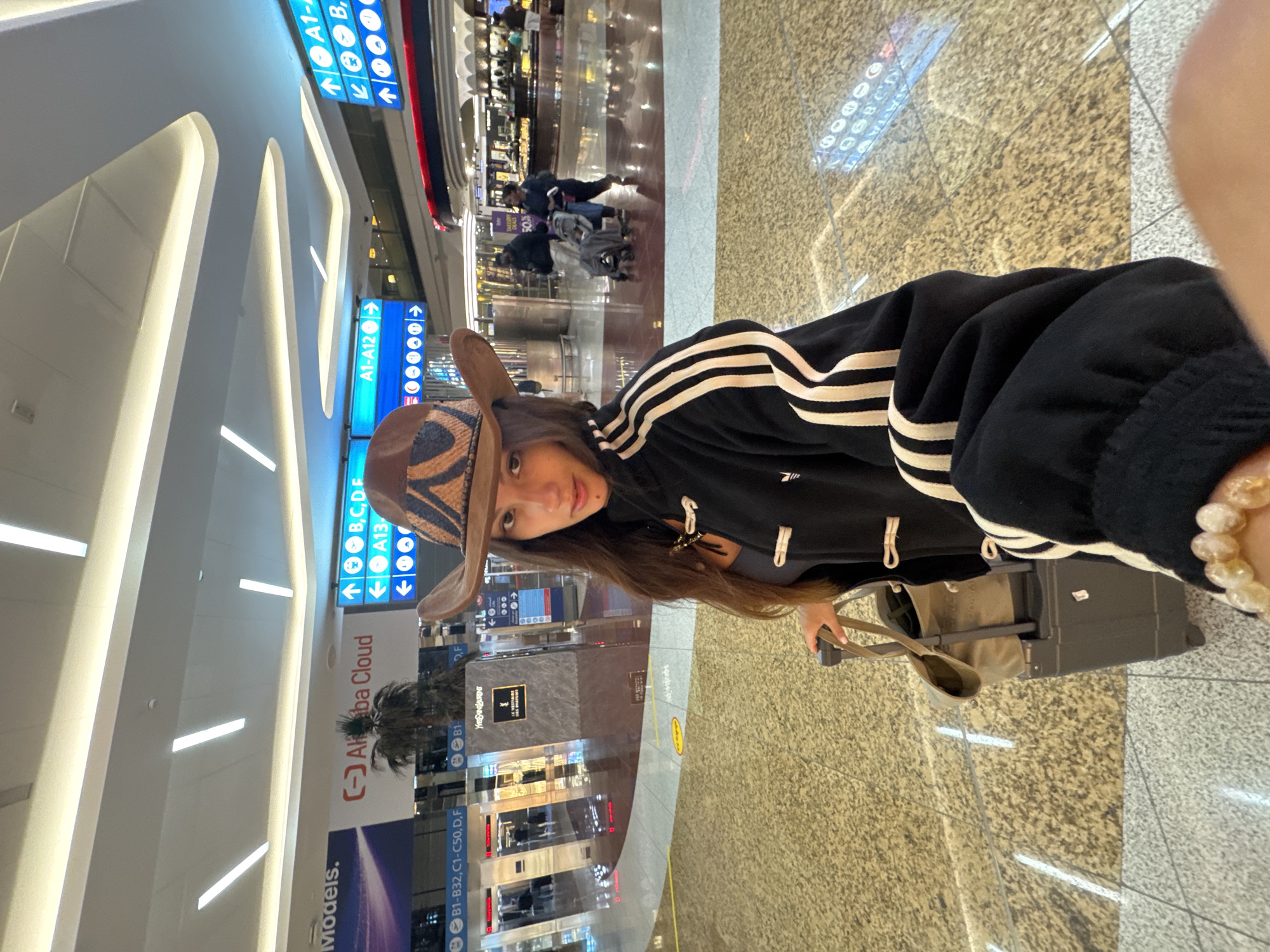 A person wearing a cowboy hat and a black Adidas jacket stands in an airport terminal. They are holding the handle of a rolling suitcase, which has a canvas tote bag on top. The background shows signs for various gates and shops, including "Alibaba Cloud" and "Yves Saint Laurent Le Vestiaire Des Parfums." People and a baby stroller are visible in the distance. The airport appears to be in Dubai, United Arab Emirates, based on the presence of an "Alibaba Cloud" advertisement, which is a prominent sponsor at Dubai International Airport. The flooring is a polished speckled pattern.