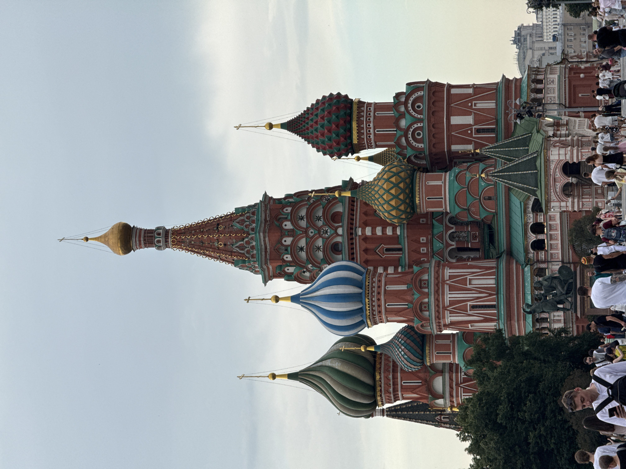 The image depicts a view of Saint Basil's Cathedral in Moscow, Russia, a famous landmark characterized by its vibrant, onion-shaped domes. The cathedral's distinctive architecture, with its red brick walls and colorful, patterned domes in shades of green, red, gold, and blue, is the primary subject. Several domes are visible, each uniquely decorated, with golden crosses atop their spires.
In the foreground and to the right of the cathedral, a crowd of people is gathered. They appear to be tourists or visitors, with many dressed casually. Some are looking at the cathedral, while others are walking or interacting with each other, suggesting a public gathering or tourist attraction. The presence of a statue in front of one of the cathedral's sections indicates a historical or memorial element.
The sky is overcast with a pale, light gray hue, suggesting it is daytime, possibly late afternoon or early morning given the soft lighting. There are no clear signs of extreme weather; the atmosphere seems calm. The overall scene conveys a sense of historical significance and popular tourism. The orientation of the image is vertical, as if the photo was taken with a phone held upright, though the content itself is captured from a slightly elevated angle looking up towards the cathedral. No visible text is present in the image.