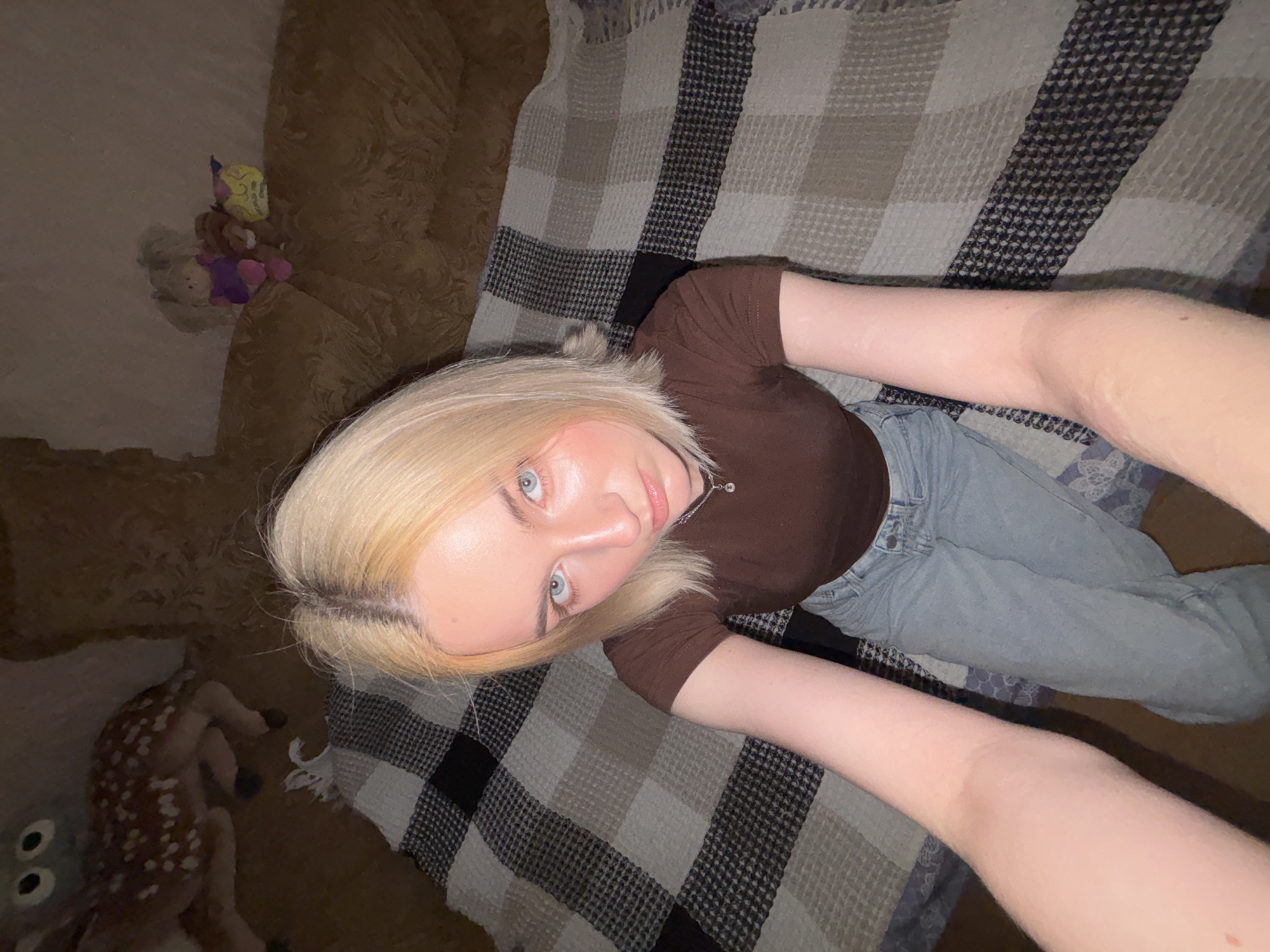 A young woman with blonde hair and blue eyes is sitting on a checkered blanket. She is wearing a brown t-shirt and blue jeans. She is looking at the camera with a neutral expression. In the background, there is a couch with several stuffed animals on it, and a stuffed deer on the floor. The scene appears to be indoors, possibly in a living room. The location is not explicitly mentioned or depicted in the image, therefore, it cannot be confirmed if it is in the city of Sinyavino, Russia.