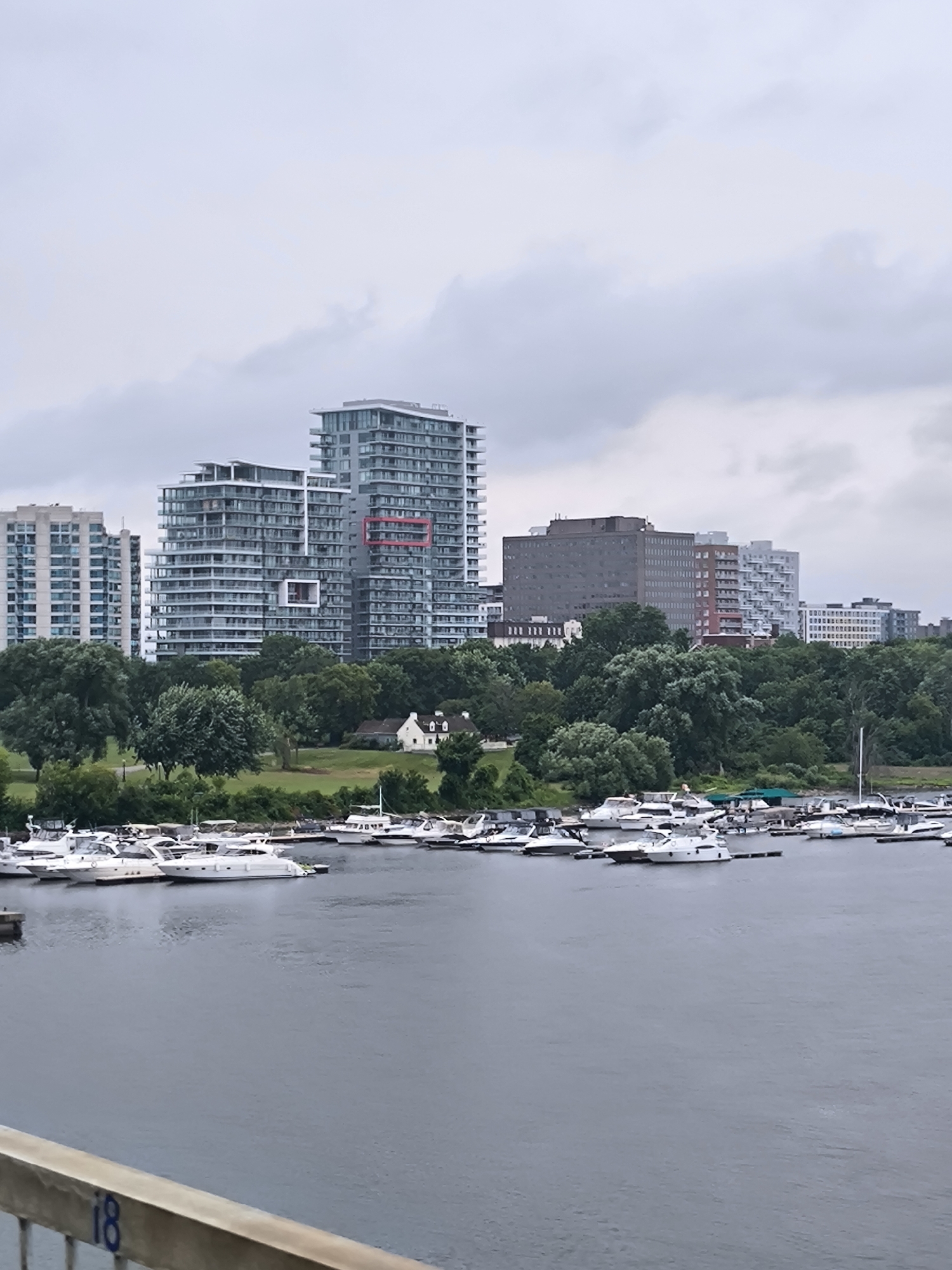 The image depicts a waterfront scene in Gatineau, Canada, under an overcast sky. In the foreground, a body of water is visible with numerous white motor yachts docked at piers. The yachts vary in size and design, with some featuring enclosed cabins and others with open decks. A wooden railing with the number "18" painted on it is present in the lower left corner of the frame.
In the midground, a lush green park area is dotted with mature trees. A small white building with a red roof, possibly a house or pavilion, is situated on the slight elevation of the park.
The background is dominated by a skyline of modern high-rise buildings. These buildings are primarily constructed of glass and concrete, with many featuring balconies. The architecture is contemporary, characterized by clean lines and multi-story structures. Several of these buildings are taller than the others, creating a varied urban silhouette. The overall impression is of a city with significant waterfront development and green spaces.