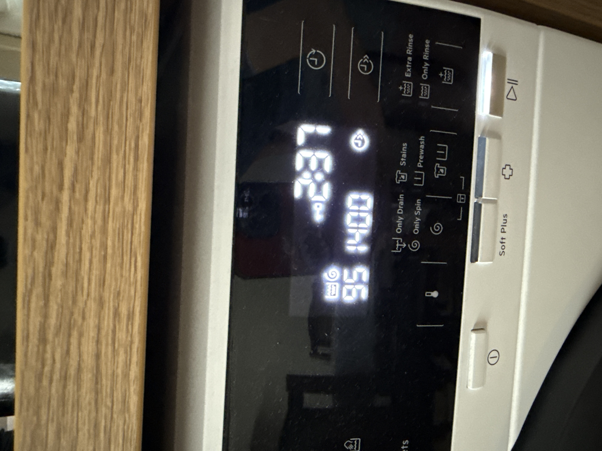 A close-up shot shows the control panel of a washing machine, displaying the timer (2:37), spin speed (1400), and temperature (95). Indicators for "Extra Rinse", "Only Rinse", "Stains", "Prewash", "Only Drain", and "Only Spin" are also visible. The 'Start/Pause' button is illuminated, along with the "Soft Plus" option. The power button is visible at the bottom. A wooden panel is on the left of the image. There is no evidence that this image was taken in Lovech, Bulgaria.