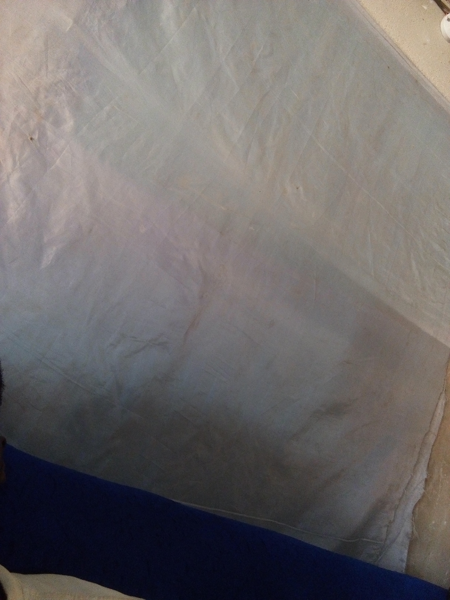 The image shows a close-up, overhead view of a crumpled, light-colored fabric, possibly a sheet or tarp. The fabric is textured with folds and creases, and there are some faint brownish stains and possibly some dirt specks visible. In the bottom left and bottom center of the image, a dark blue fabric is visible, partially obscuring the light-colored fabric. The scene appears to be indoors, possibly a domestic setting. There is no identifiable location in the city of Nyabe, Nigeria, visible in this image.