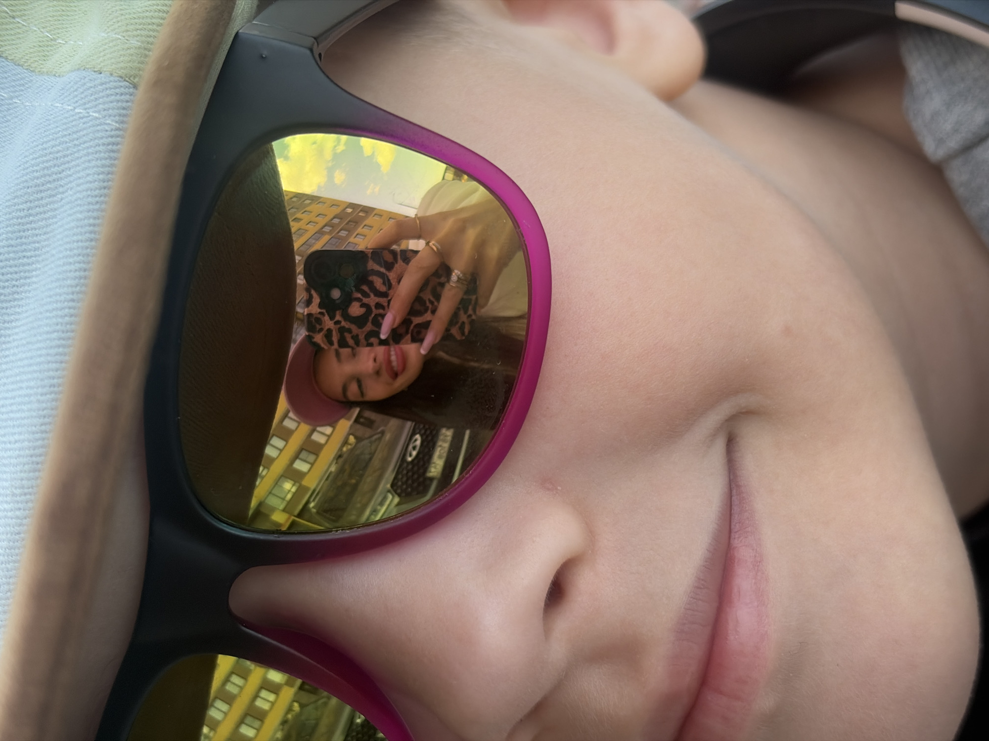 The image presents a close-up of a child's face, likely an infant or toddler, with fair skin and a subtle, gentle smile. The child is wearing black sunglasses with fuchsia/purple accents on the frames. A light-colored fabric, possibly a hat, is visible on the left side of the frame, and a grey garment on the right.
The mirrored, iridescent yellow-gold lenses of the sunglasses reflect an inverse scene. Prominently featured is an adult, likely female, holding a smartphone with a distinctive leopard-print case. Her hands, adorned with rings and manicured nails, are visible, and she appears to be wearing a light-colored top and possibly a pink hat or headband. Her face, reflected upside down, also shows a smile.
Behind the reflected adult, an urban environment is visible. This includes a multi-story building with numerous windows, suggesting a city structure, and part of a dark car's grill and a blurry license plate in the lower portion of the reflection. The reflection also captures a bright sky with clouds. The scene is illuminated as if by daylight. The photograph captures a candid, cheerful moment, potentially a selfie or a photo taken of the child, with the photographer's reflection in the sunglasses. The context suggests the location is Saint Petersburg, Russia. No clear, legible text is visible.