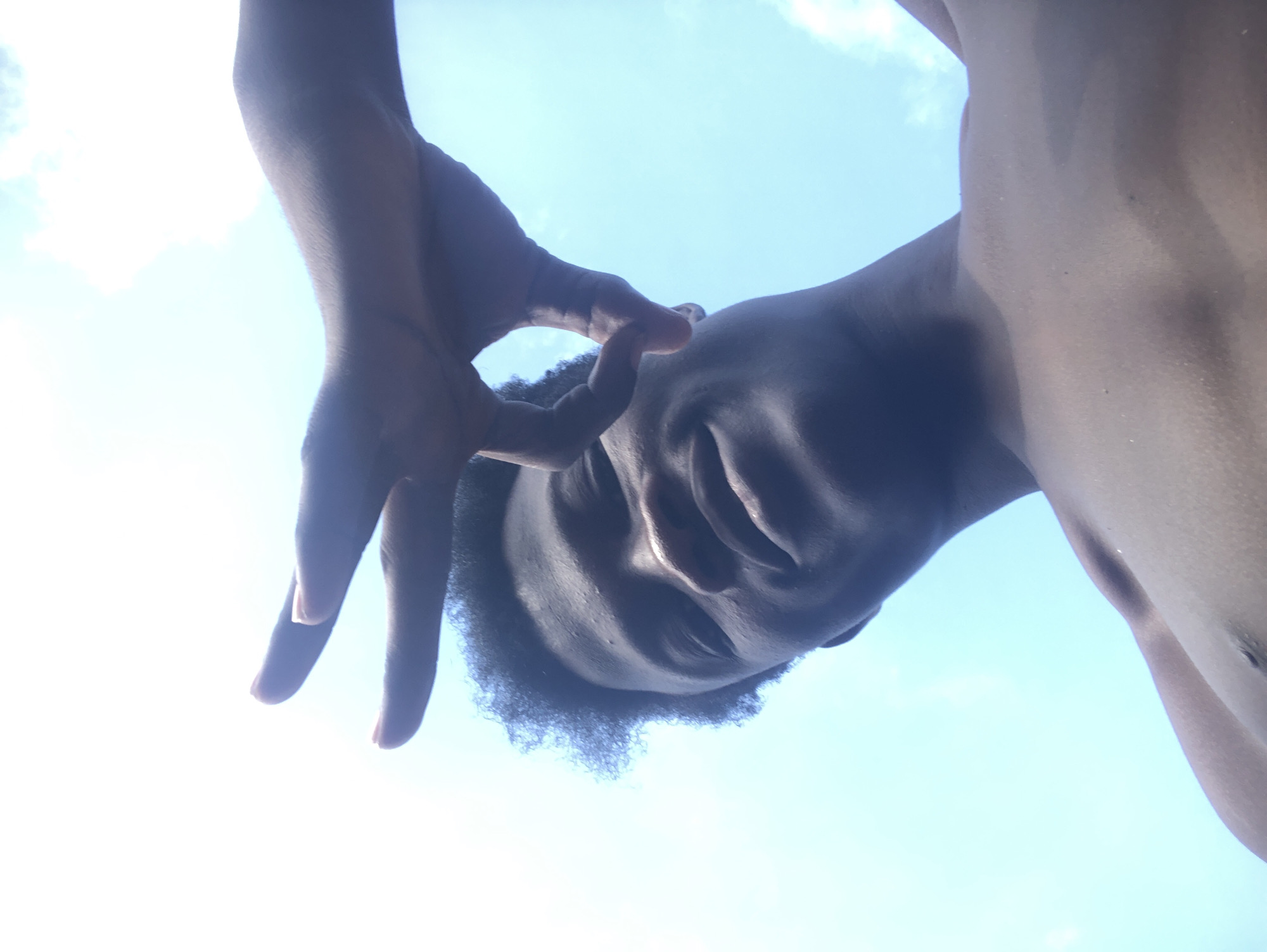 A low-angle, close-up shot features a young Black male outdoors with a bright sky as the background. His head is tilted back, and his face is partially obscured by his hand, which is raised to cover his eye. His hair is short and dark, and his skin is dark-skinned. The man appears to be shirtless, revealing his chest and shoulders. The sky is a light blue with some wispy white clouds, suggesting it is daytime. The overall impression is a casual, candid moment captured outdoors in Ado, Nigeria.