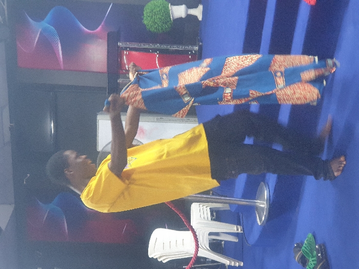 An indoor scene features a barefoot male subject, dressed in a yellow short-sleeved t-shirt and dark trousers, standing on a blue carpeted floor. He holds a length of blue, orange, and brown patterned fabric with both hands, extended forward and downwards. His head is turned to the right, with a slight smile. To the left, a stack of white plastic chairs is situated behind a red rope barrier connected to a chrome stanchion. A pair of green and black sandals rests on the blue carpet near the subject's left foot. To the right, a blue carpeted raised platform with visible steps supports a dark podium and a white urn containing a spherical green artificial plant. The background wall is dark with two large, abstract graphic designs featuring swirling lines in blue, pink, and red. The specific location within Onitsha, Nigeria, cannot be determined from the image.