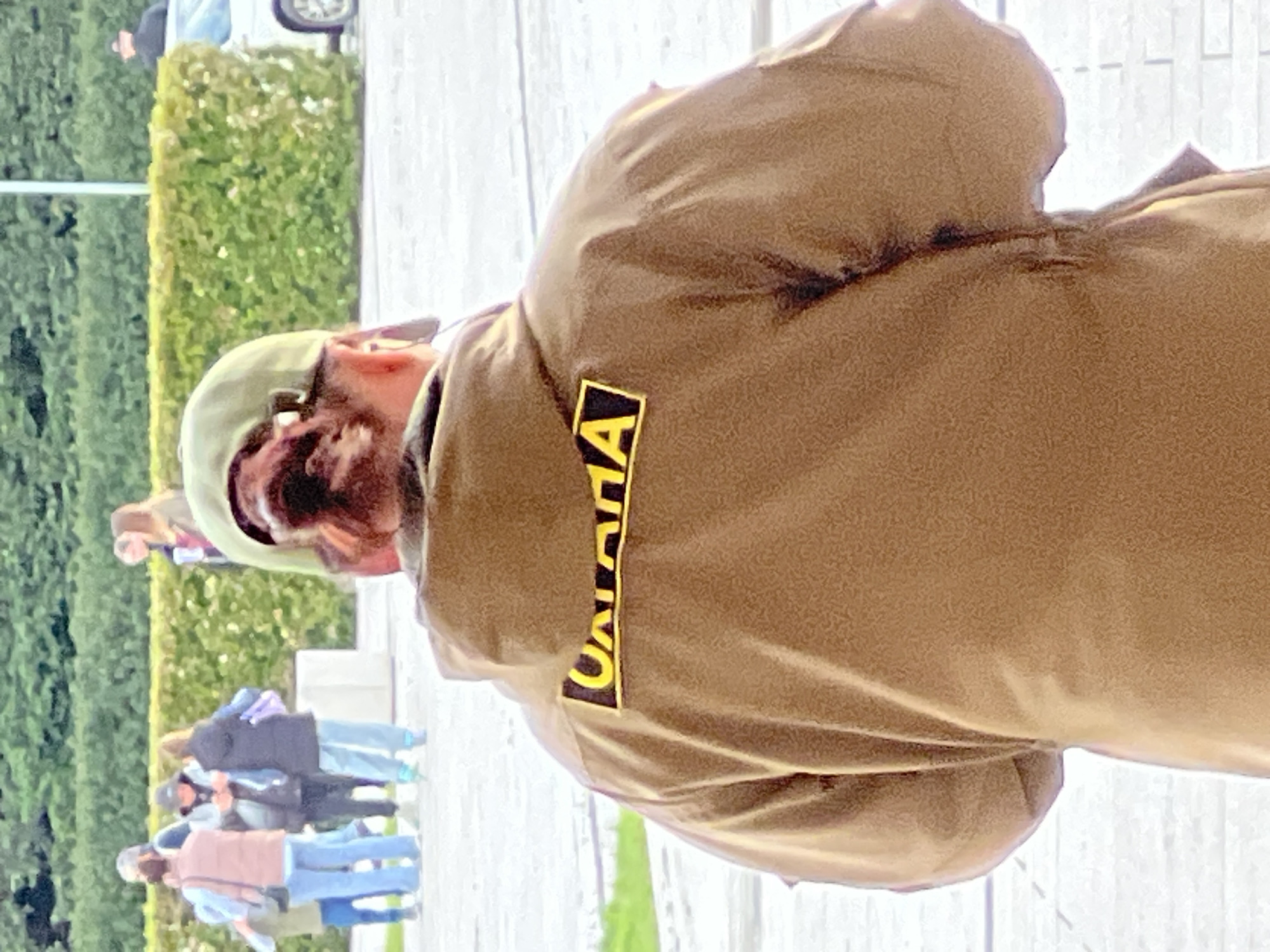 The image, when viewed in its correct vertical orientation, presents an outdoor scene in what appears to be a public park or spacious promenade in Krasnodar, Russia, bathed in bright daylight.
The central subject is a person viewed from behind and slightly to their right, wearing a light brown, possibly olive-drab, jacket or shirt. They are also wearing a light-colored baseball cap, beneath which dark hair and a beard are partially visible. Sunglasses are resting on the cap or the back of their head. A prominent detail is a black rectangular patch with bold yellow letters on the upper back of their jacket, which clearly spells out "MONTANA".
In the middle ground and background, several other people are visible, indicating a public gathering or common thoroughfare. These individuals are casually dressed and are seen walking or standing on a light-colored paved surface. Green, well-maintained hedges and trees line the paved area, suggesting a landscaped environment. Further in the distance, a portion of a white car is visible, and one person appears to be pushing a stroller or a small cart. The overall impression is one of a calm, everyday public space on a clear, bright day.