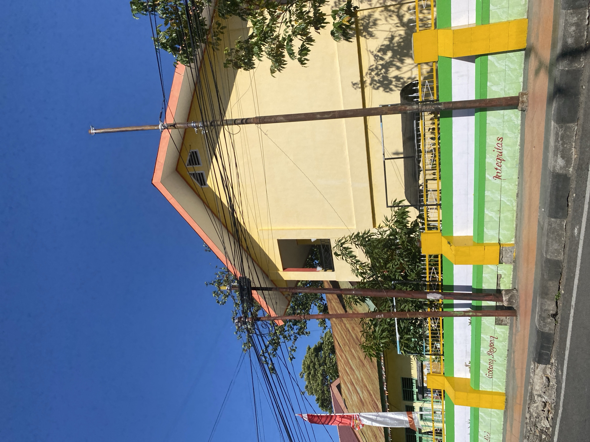 The image depicts a street view in Bantaeng, Indonesia, on a clear, sunny day. A yellow building with an orange roofline dominates the background, its facade casting shadows on the ground. In front of the building, a colorful wall with green and white stripes, accented by bright yellow pillars, serves as a barrier. The words "Gotong Royong" and "Integritas" are visible, written in cursive on the lower section of the wall. A yellow fence runs along the top of this wall, behind which a black canopy structure is partially visible. Several utility poles with a complex network of black wires crisscross the scene. A red and white Indonesian flag is affixed to a pole on the left side of the frame. Trees with green foliage are scattered throughout the scene, some encroaching on the building and fence. The foreground shows a paved road and a concrete curb, suggesting an urban or semi-urban setting. There are no people clearly visible in the image.