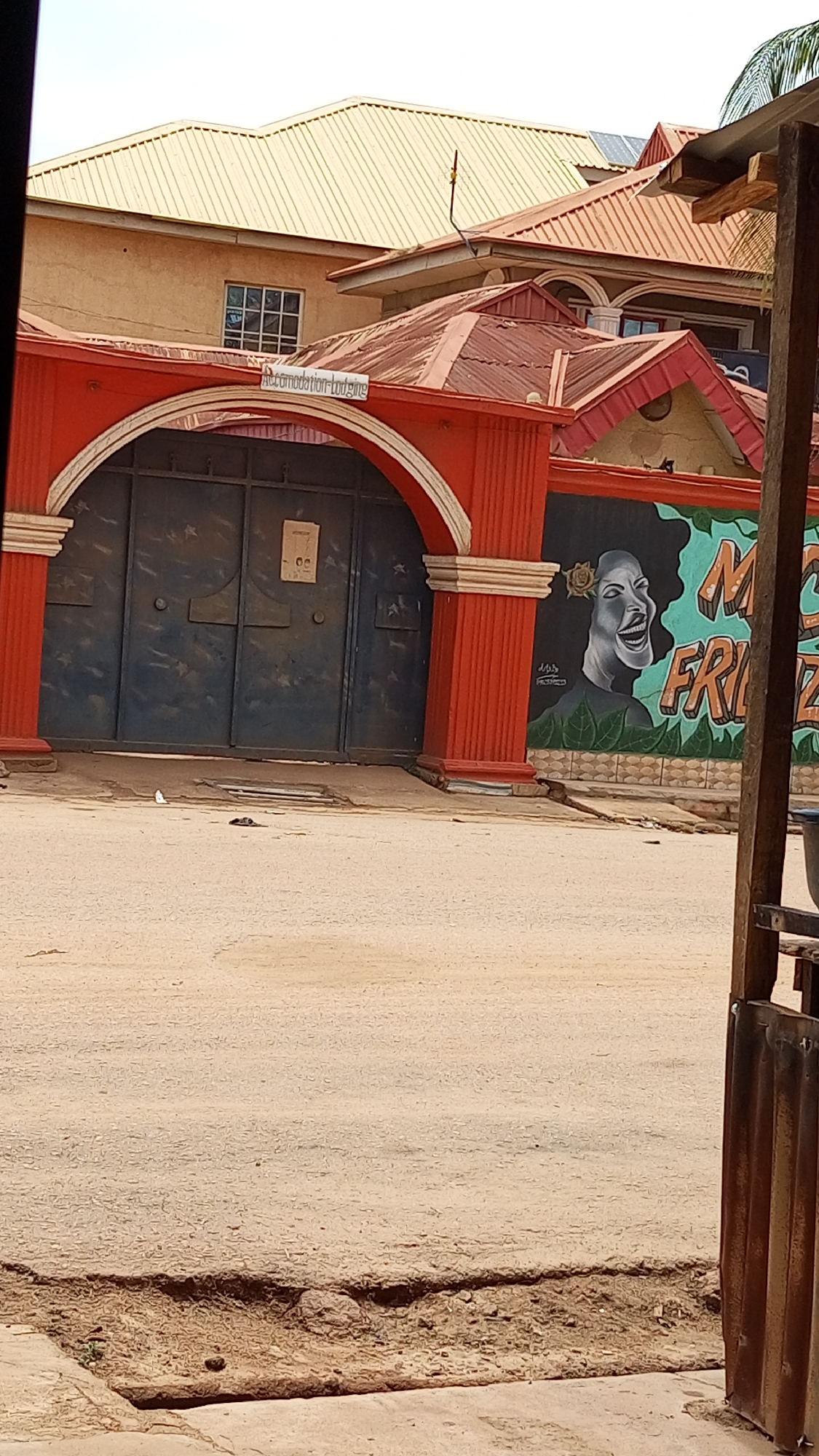 This image captures a street view in Sabon Tasha, Nigeria, featuring the entrance to a property. In the foreground, a light brown, unpaved or gravely road shows some unevenness and a small ditch-like area.
The central focus is a large, dark blue/grey metallic double gate set within a prominent red arched frame. The arch is supported by red columns with white base and capital details. Above the gate, a white rectangular sign clearly displays the text "Accommodation-Lodging."
To the right of the gate, a wall is adorned with a vibrant mural. The mural features a stylized, black and white portrait of a person (likely a woman) with a wide, joyful smile or laugh, and a yellow flower in her hair. This portrait is set against a teal-colored background with green leaf motifs at the bottom. Partial text on the mural is visible, reading "M" and "FRIENDZ," possibly part of a phrase like "My Friendz." Below the portrait, a faint signature resembling "Artist D'Rapp" can be discerned.
Behind the gate and wall, parts of several buildings are visible. One building has a light brown/tan facade and a window with white grilles. Another features a reddish-brown corrugated metal roof, while a larger structure in the background boasts a yellowish/golden corrugated metal roof. Solar panels are faintly visible on one of the roofs.
The scene takes place during the daytime under a clear sky, indicated by the bright and even lighting. There are no people present, apart from the depiction in the mural. The overall impression is a common streetscape in a Nigerian residential or commercial area.